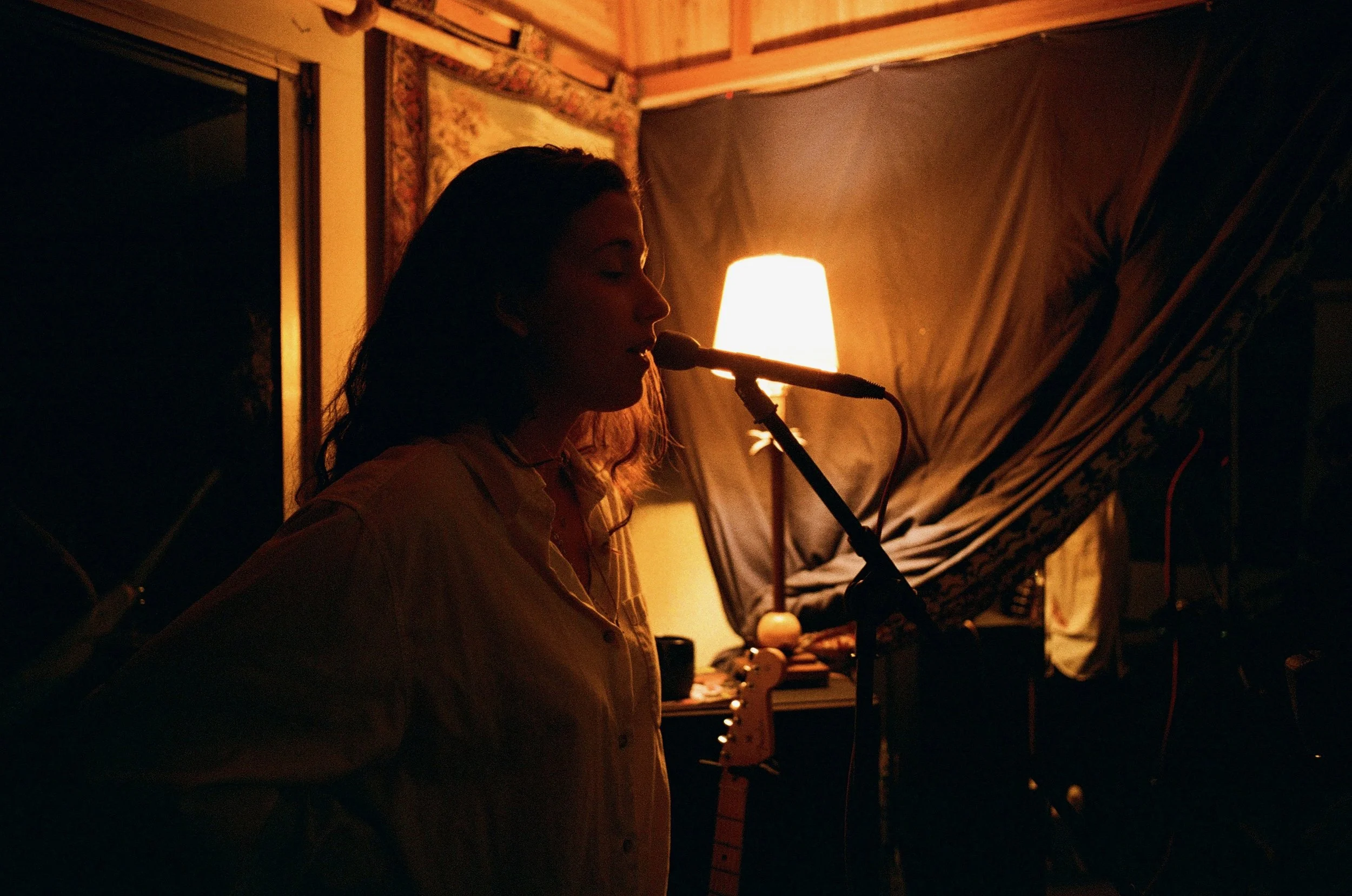 A person with long dark hair singing into a microphone in a dimly lit room illuminated by a warm lamp.
