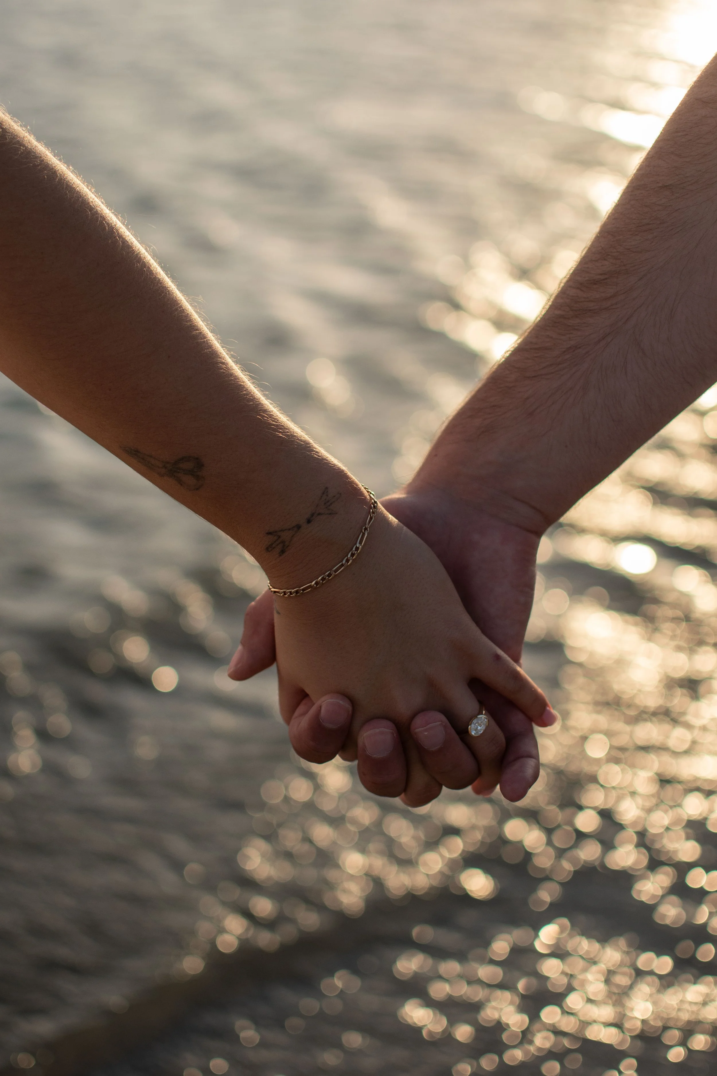 Two people holding hands over water during sunset, with sunlight reflecting on the water's surface.