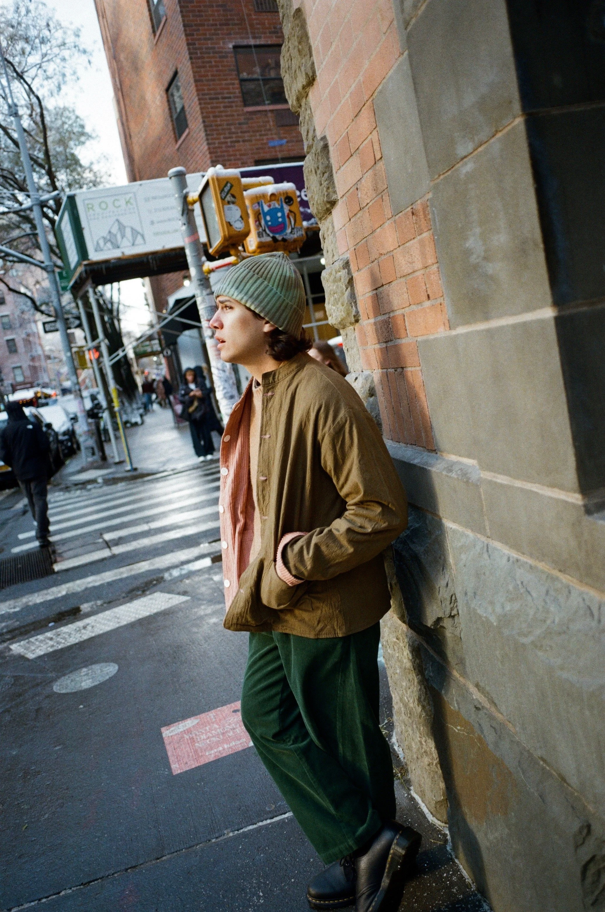 A person standing on a city sidewalk, leaning against a brick and stone building wall, wearing a green beanie, a brown jacket, green pants, and black boots. There are pedestrians and storefronts visible across the street, with some street signs and t