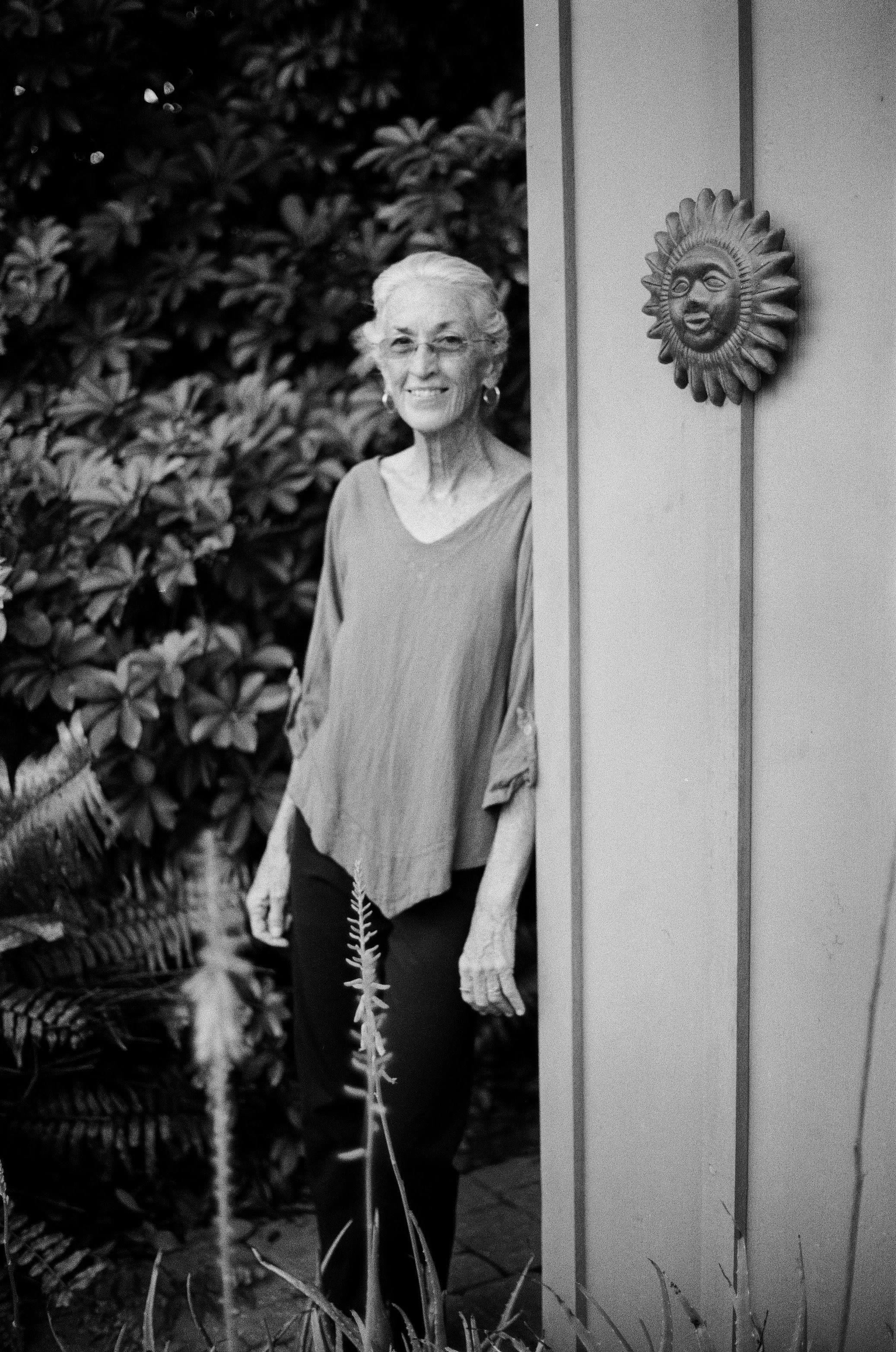 An elderly woman with glasses and earrings is standing outdoors next to a wall with a decorative sun face sculpture, and there are lush plants behind her.