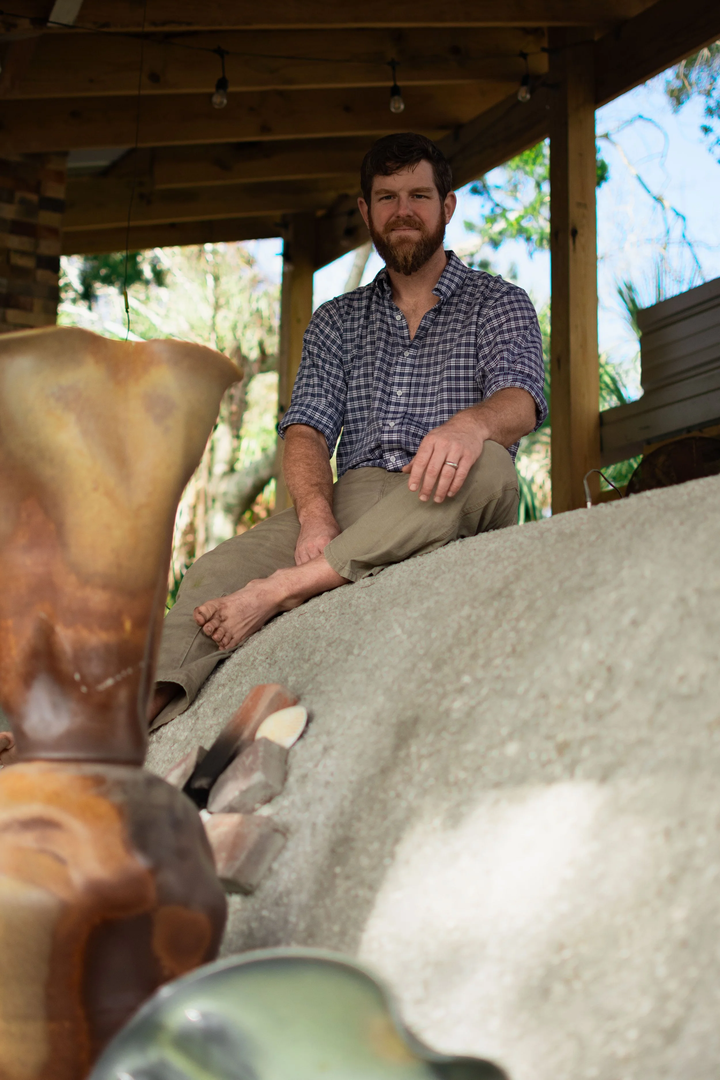 A man with a beard and short dark hair sitting on a carpeted floor outside, underneath a wooden porch with string lights, wearing a checkered shirt and khaki pants, with trees and blue sky in the background.
