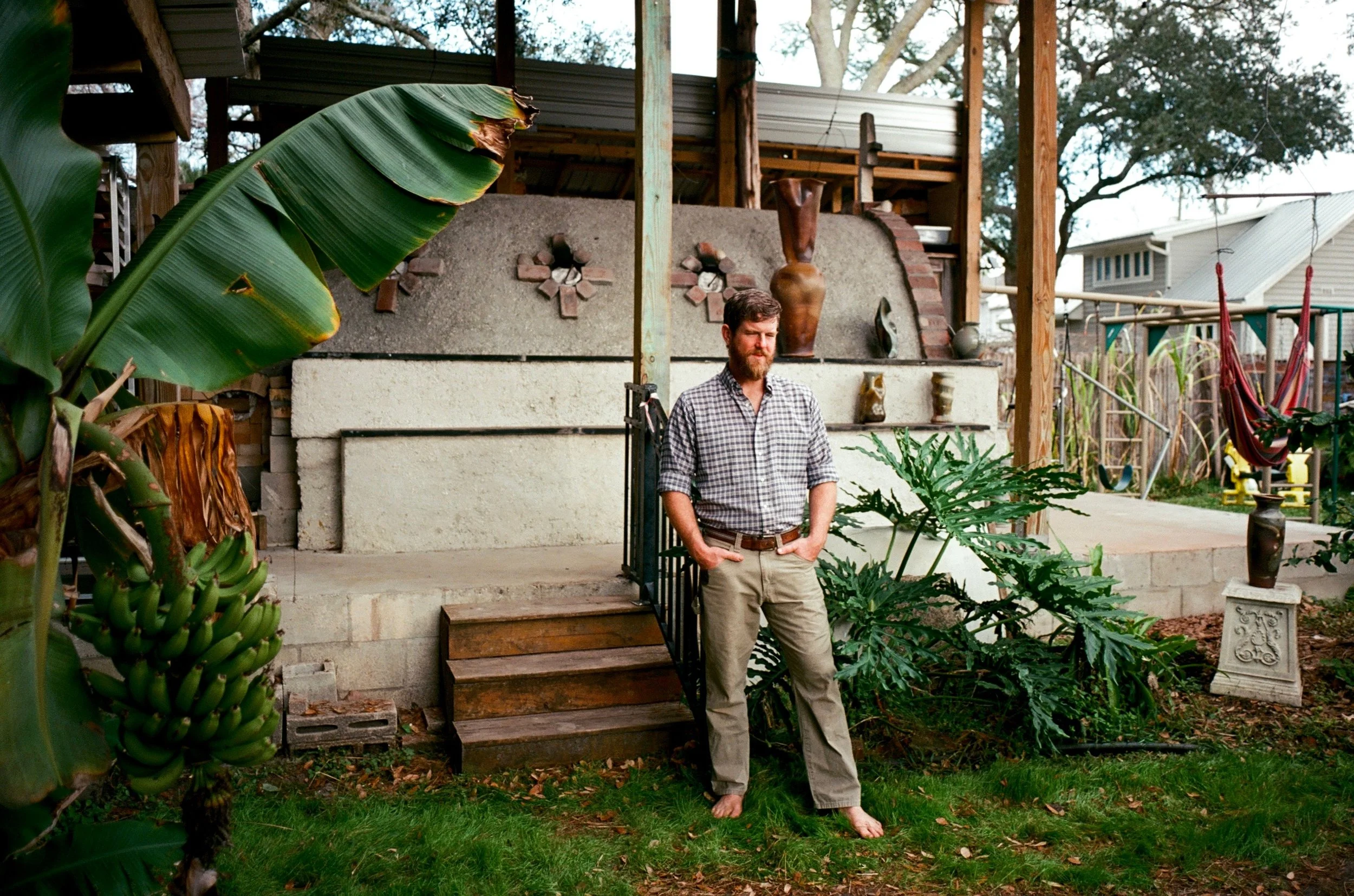 A man with a beard, wearing a checkered shirt and khaki pants, stands outdoors on a patio with his hands in his pockets, barefoot. There are lush green plants, including a banana plant with a bunch of bananas, surrounding him. In the background, ther