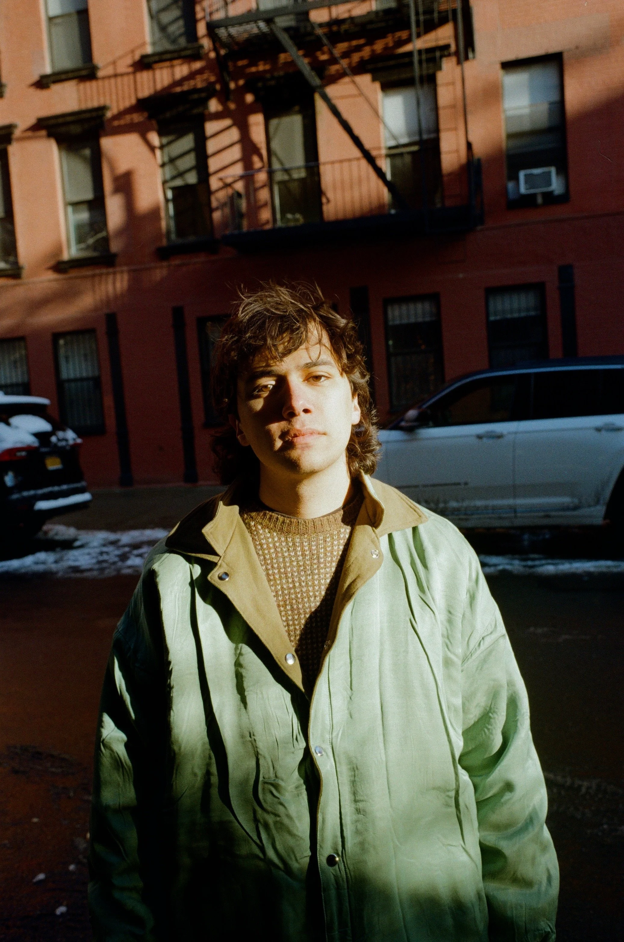 A young man with wavy hair standing outdoors in front of a red brick building with black fire escape stairs. He is wearing a tan jacket over a patterned sweater, and sunlight illuminates his face.