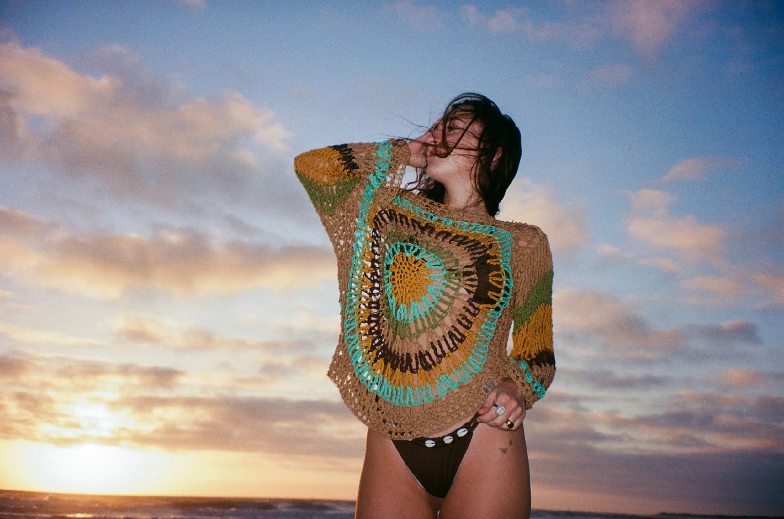 A woman standing on the beach at sunset, wearing a colorful crocheted top and black bikini bottom, with her hair blowing in the wind.