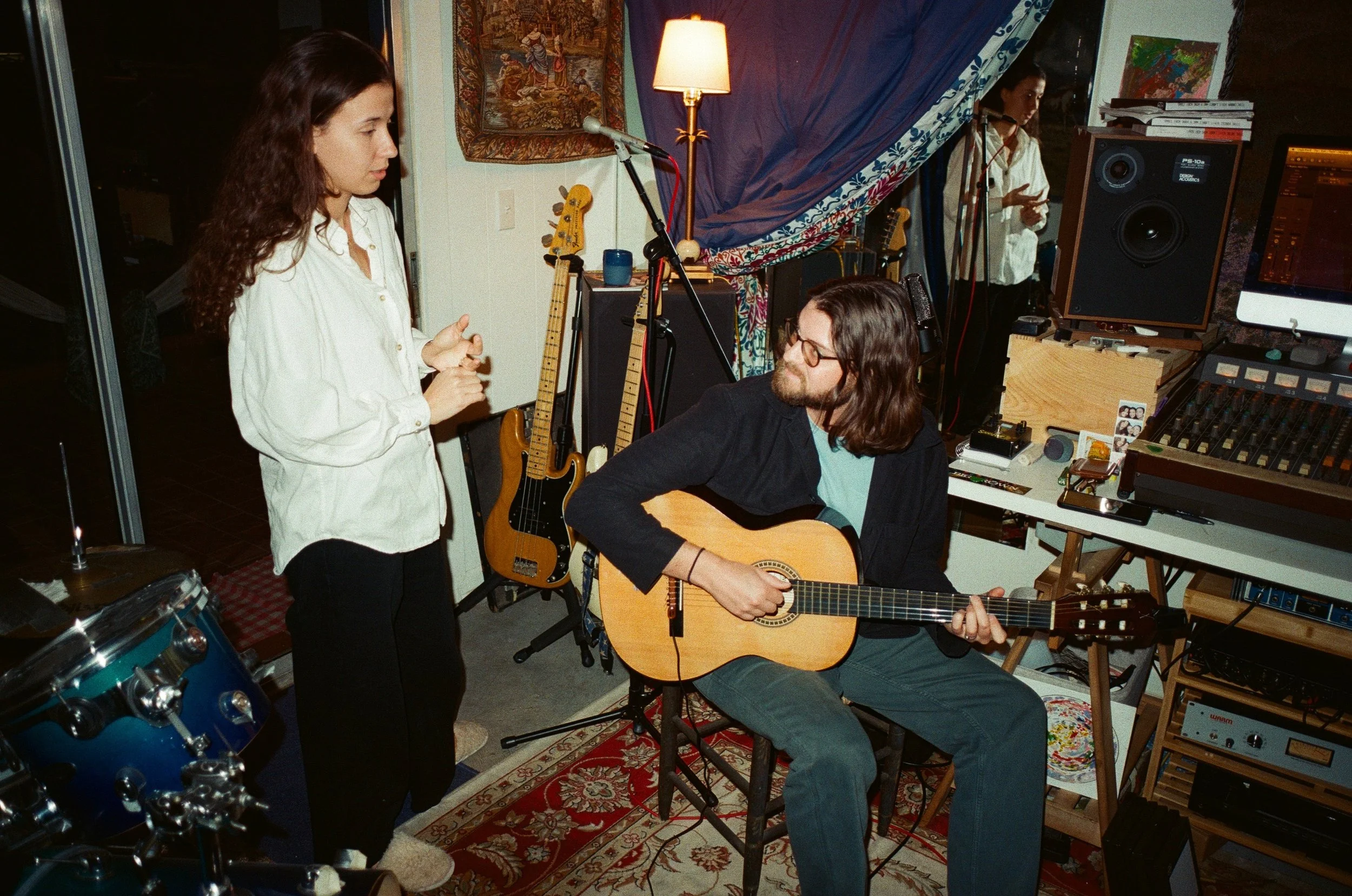 Two musicians in a cozy room with musical instruments and recording equipment. A woman with long brown hair and glasses is playing an acoustic guitar, while a woman with long curly brown hair in a white shirt is standing nearby, possibly singing or s