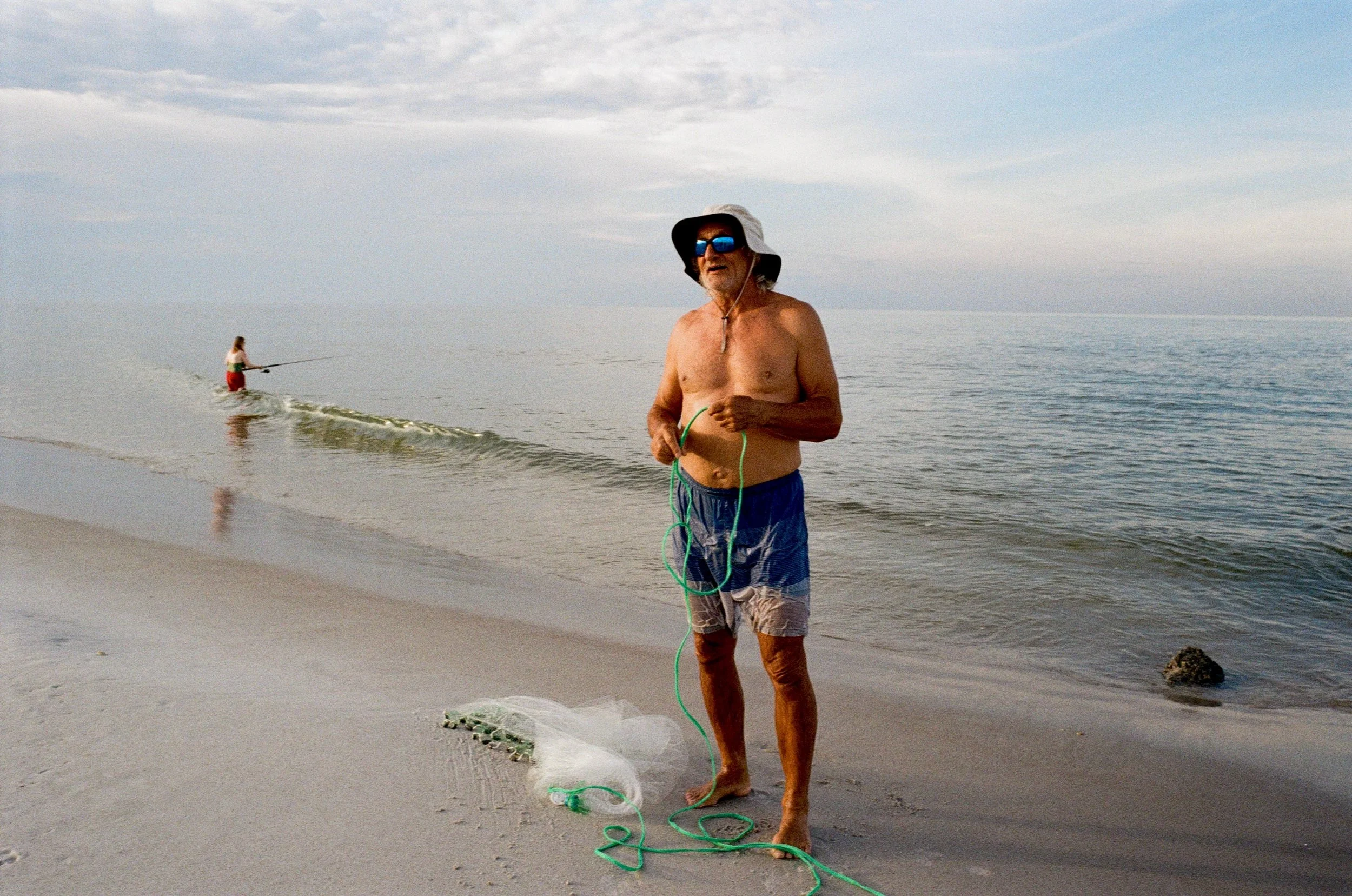 An elderly man standing at the edge of the beach, holding a fishing net, with a younger person fishing in the water in the background.