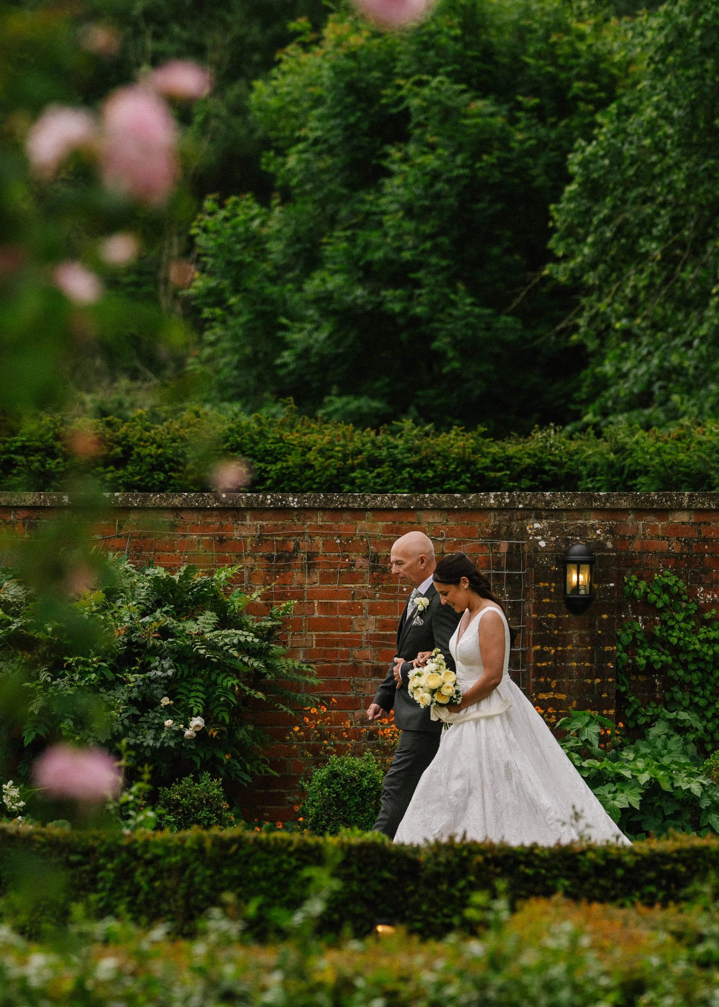 A bride and groom walking outdoors in a garden, with a brick wall and lush greenery in the background, overlooking a hedge and pink flowers in the foreground.