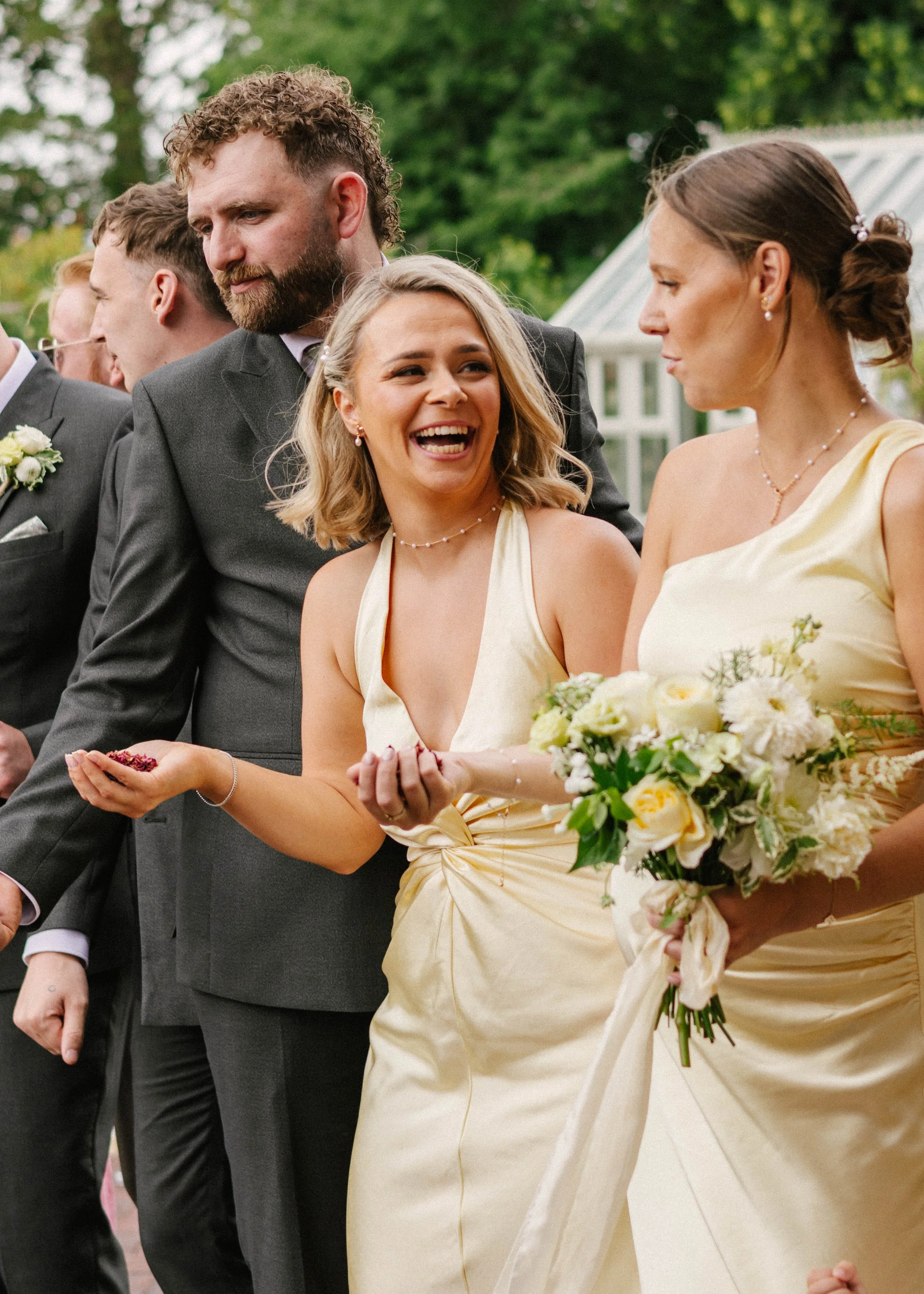 A group of people dressed for a wedding, including a woman in a yellow dress holding a bouquet of white flowers, smiling and talking with others outdoors with green trees in the background.