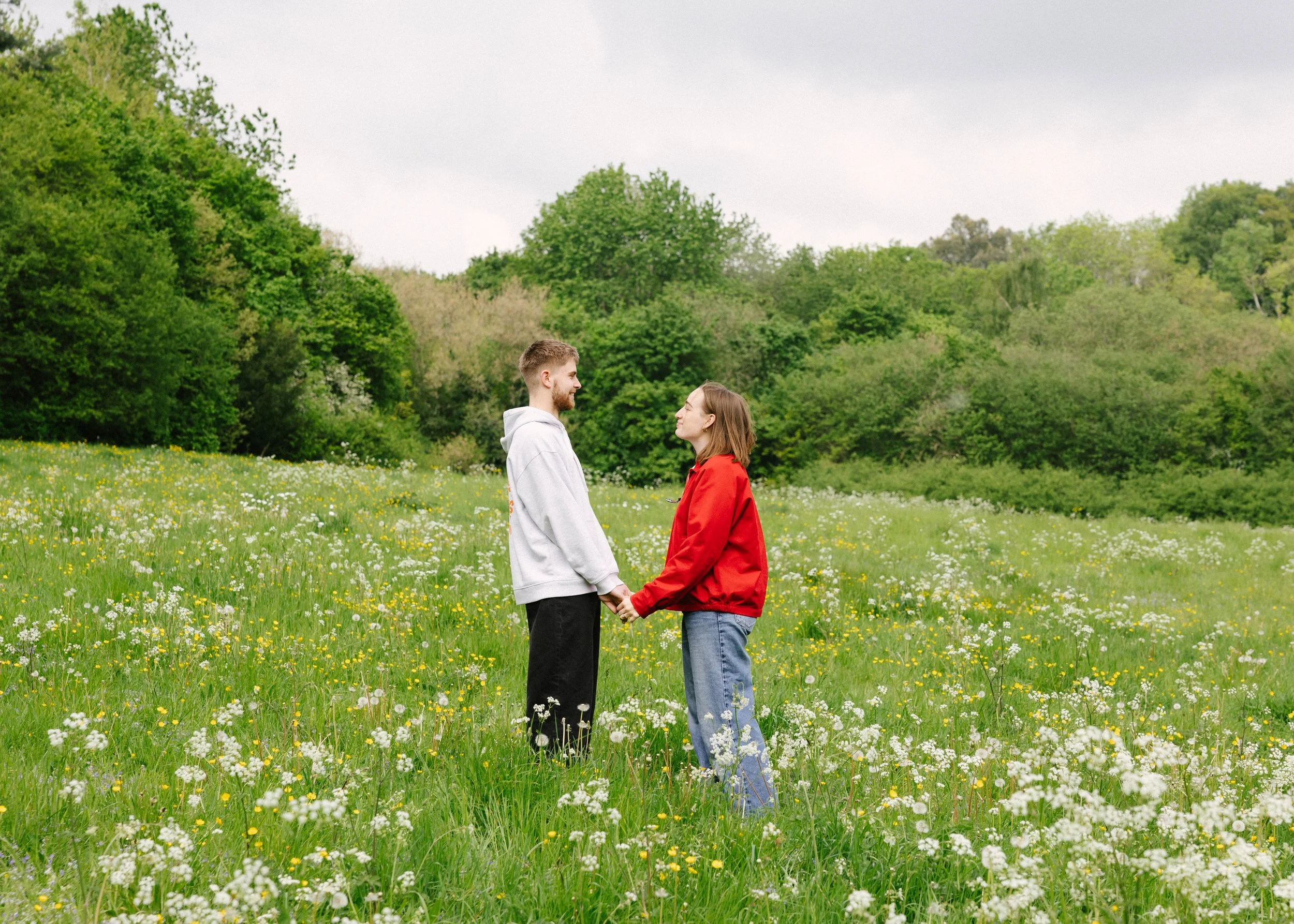 A young man and woman holding hands in a field of wildflowers, facing each other and smiling, with trees and an overcast sky in the background.
