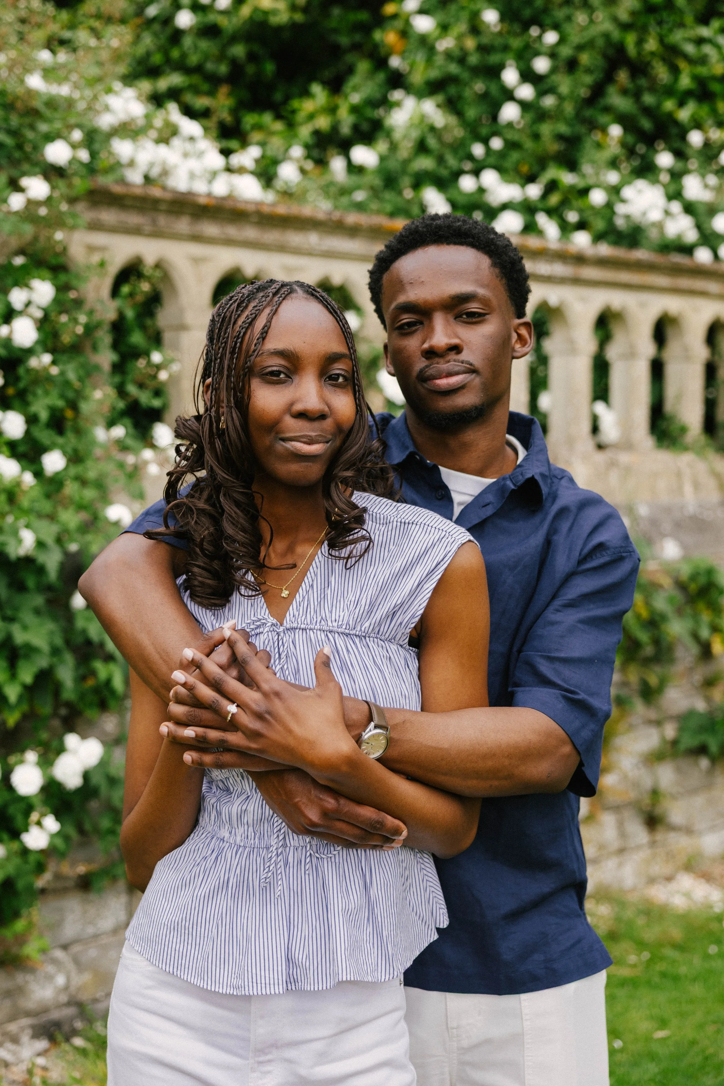 A young couple hugging outdoors in front of a stone balustrade with greenery and white flowers behind them. The woman has dark skin and braided hair, and is wearing a sleeveless striped top. The man has dark skin and short afro hair, and is wearing a