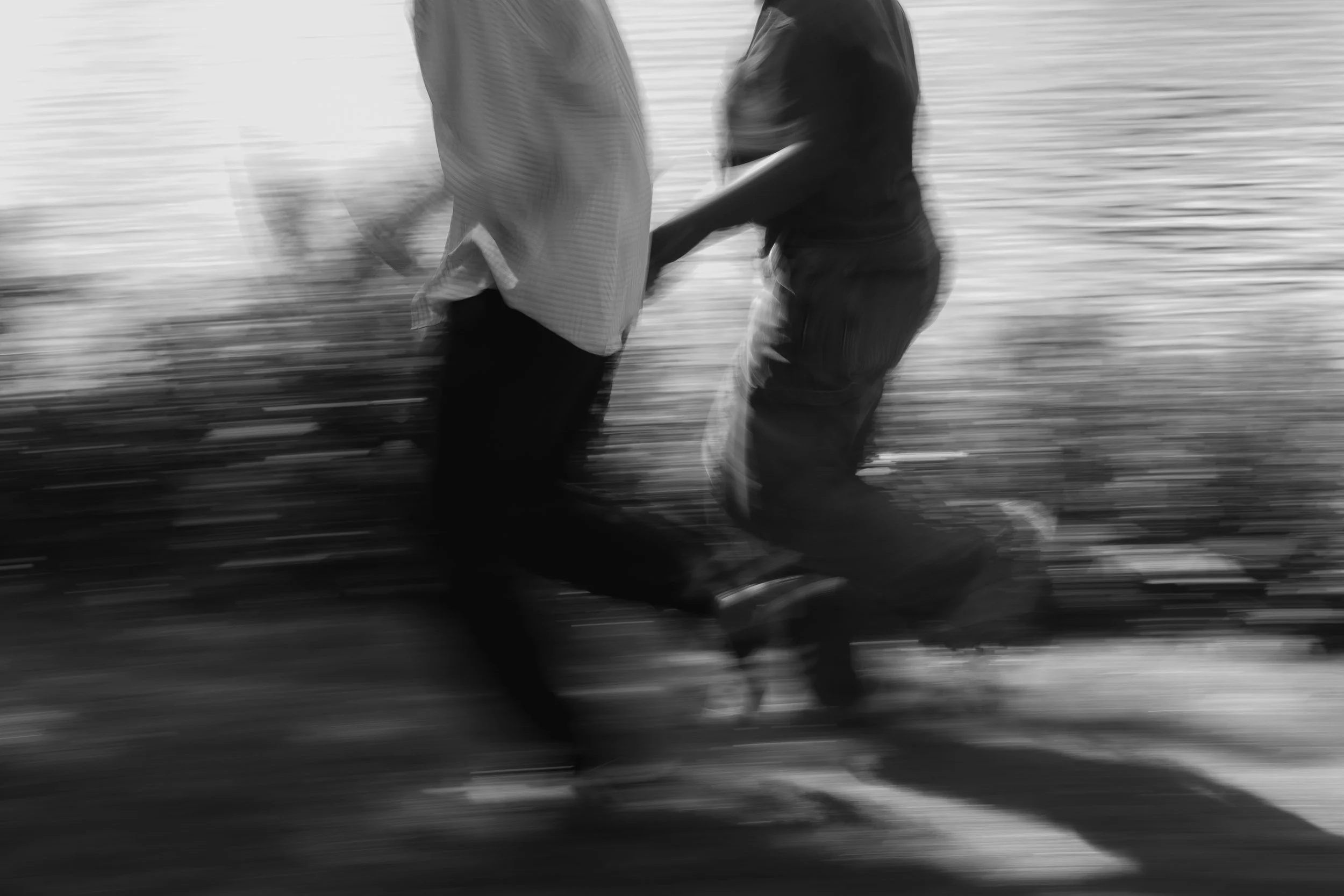 Black and white photo of two people holding hands while walking near a body of water, with a blurred background.
