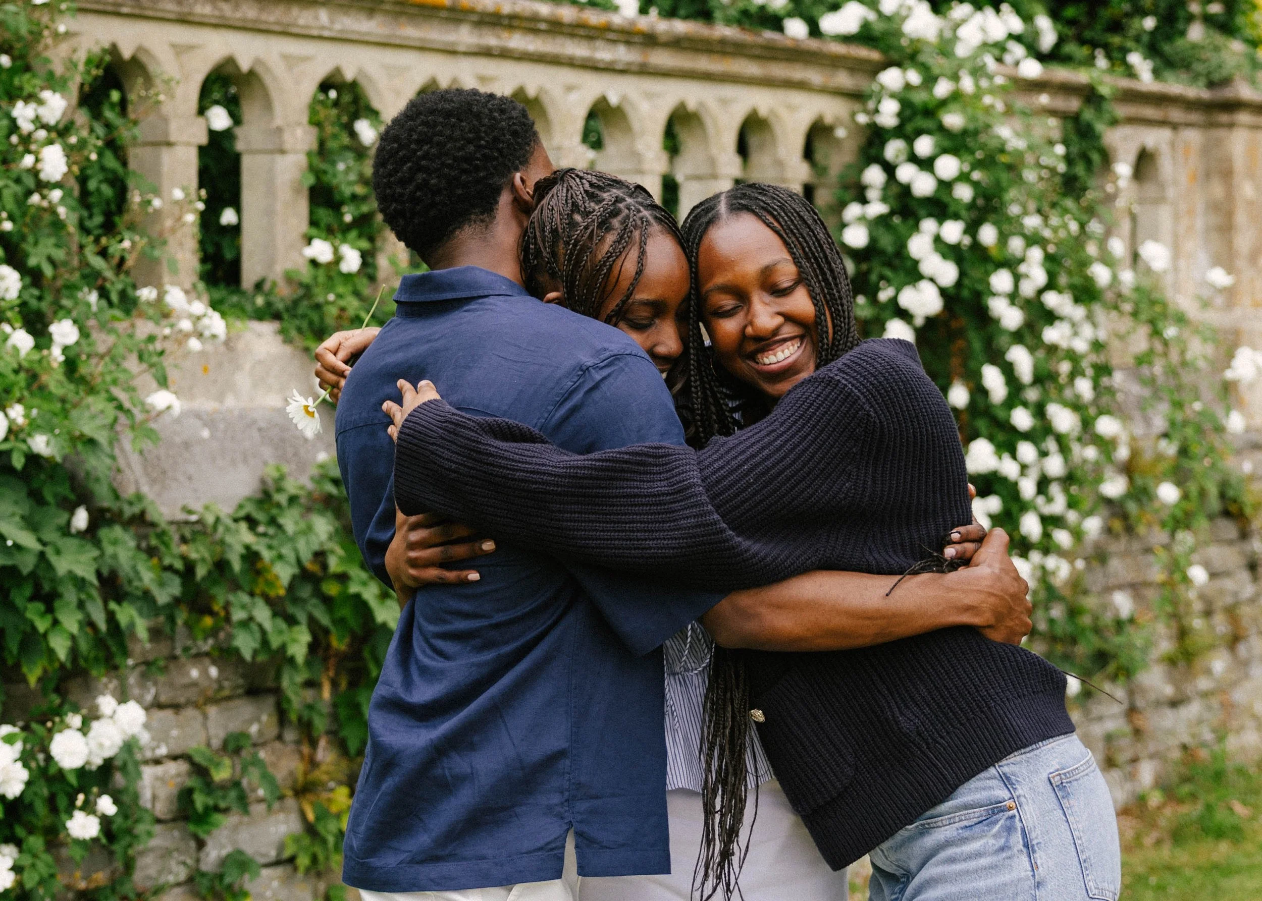 Three people hugging each other outdoors near a stone wall with greenery and white flowers.