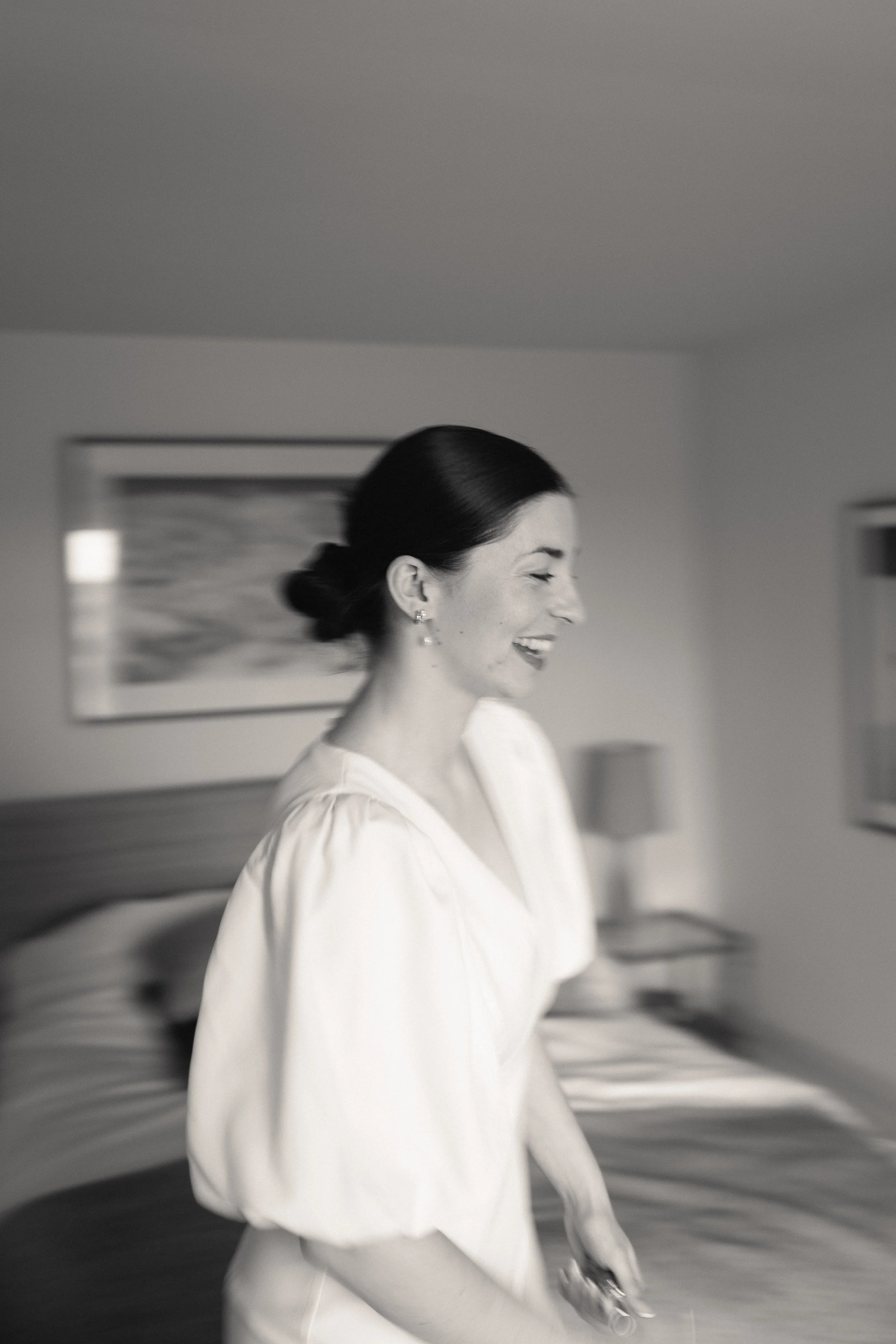 A woman with dark hair tied in a bun, smiling while walking indoors in a bright room with a painting and furniture in the background.