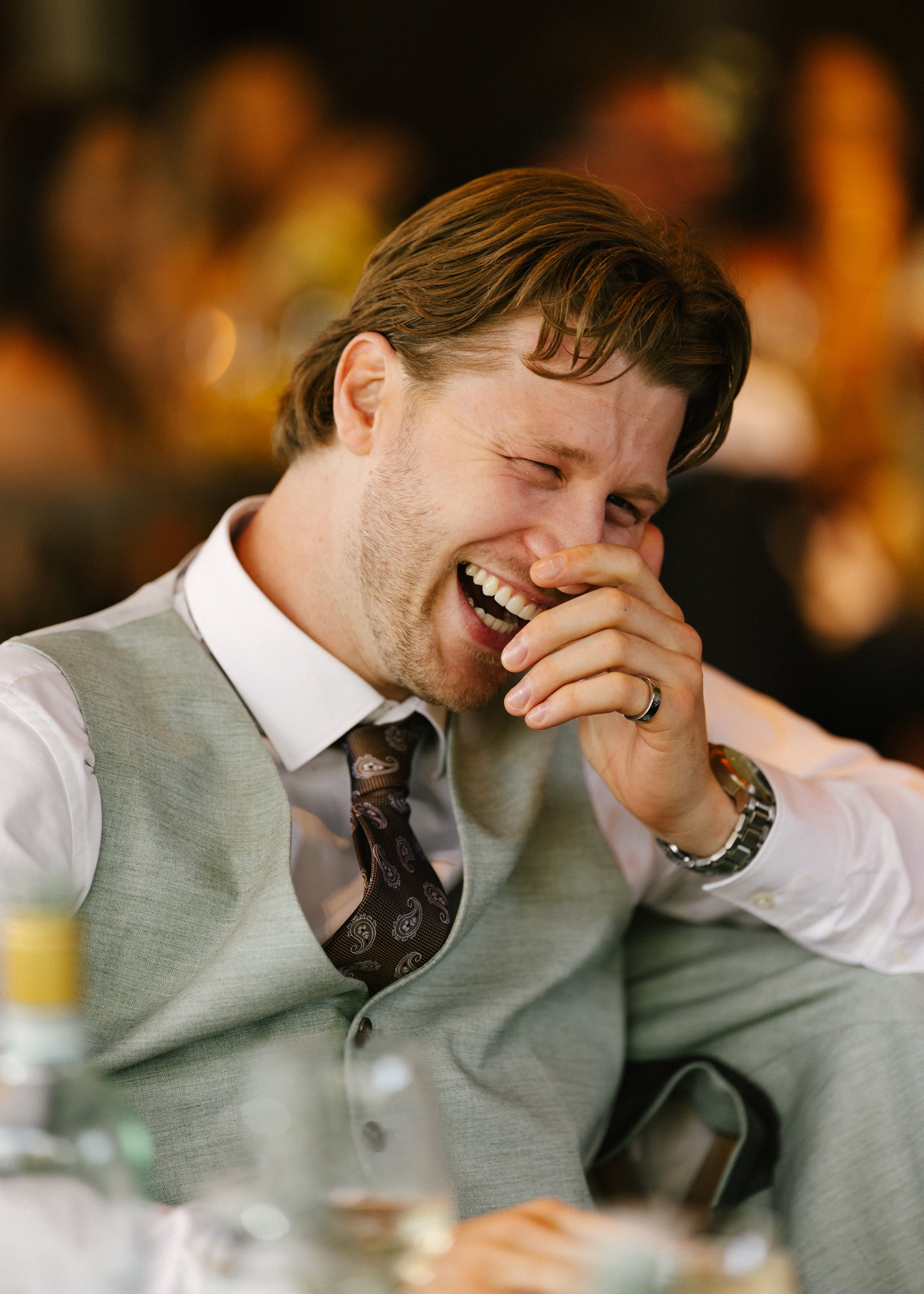 A man with brown hair, wearing a gray vest and a white shirt, is laughing with his eyes closed and his hand covering his mouth. He is wearing a wedding ring and a watch. The background is blurred with warm lighting.