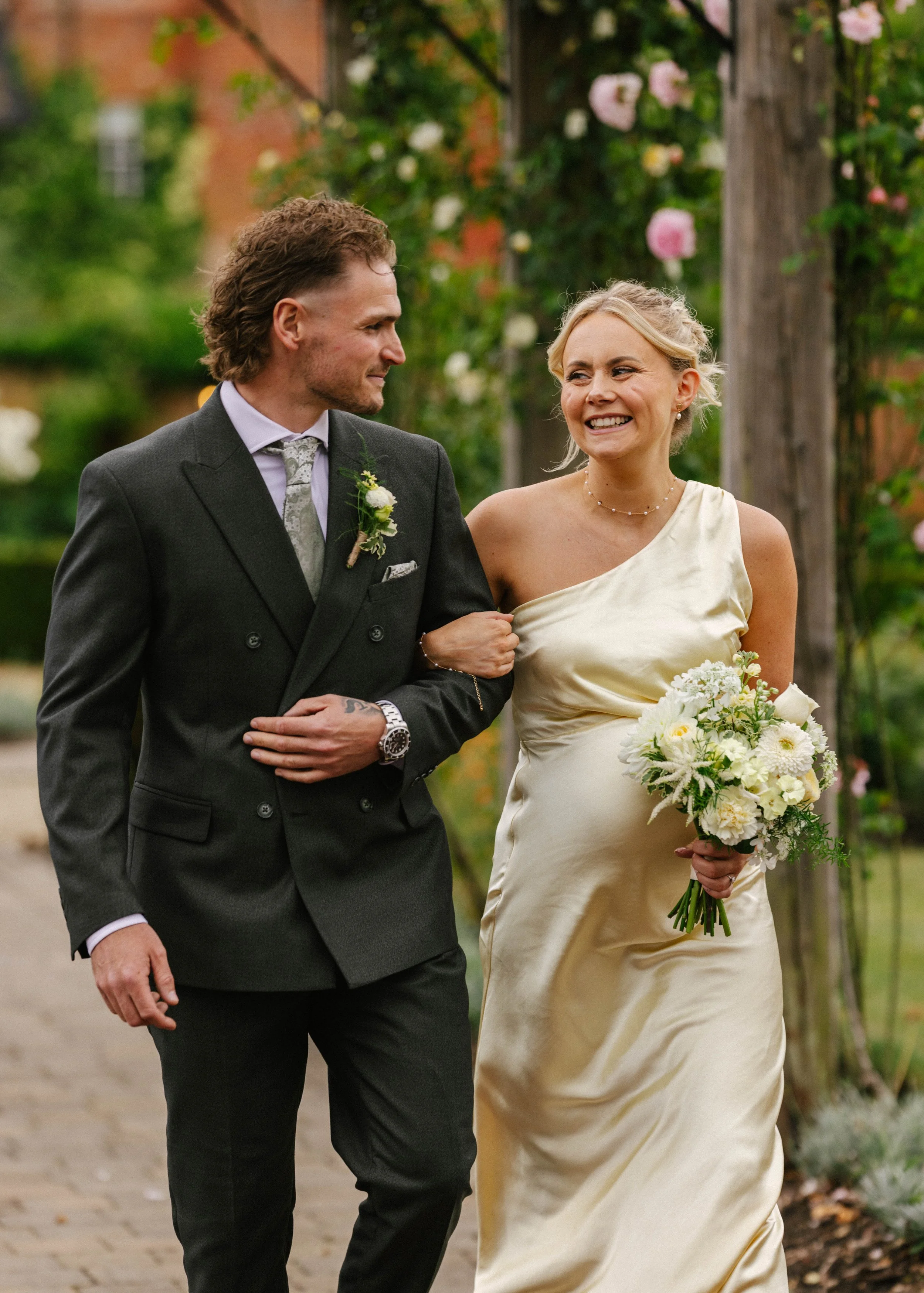 A man and woman walking arm-in-arm outdoors during a wedding, with the woman smiling and holding a bouquet of white flowers, and the man dressed in a dark suit with a boutonnière, in front of greenery and blooming flowers.