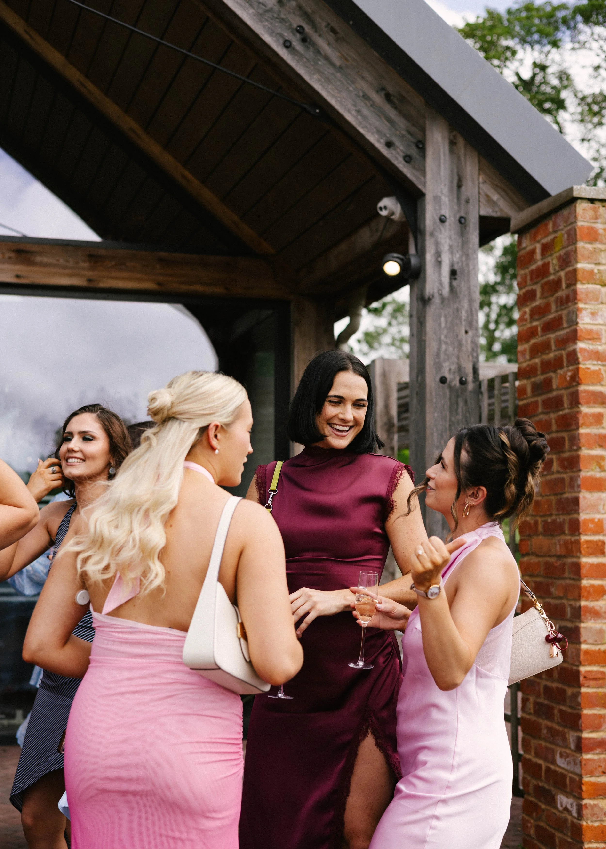 A group of women at an outdoor gathering, dressed in colorful dresses, holding champagne glasses, and enjoying conversation under a wooden and brick structure.