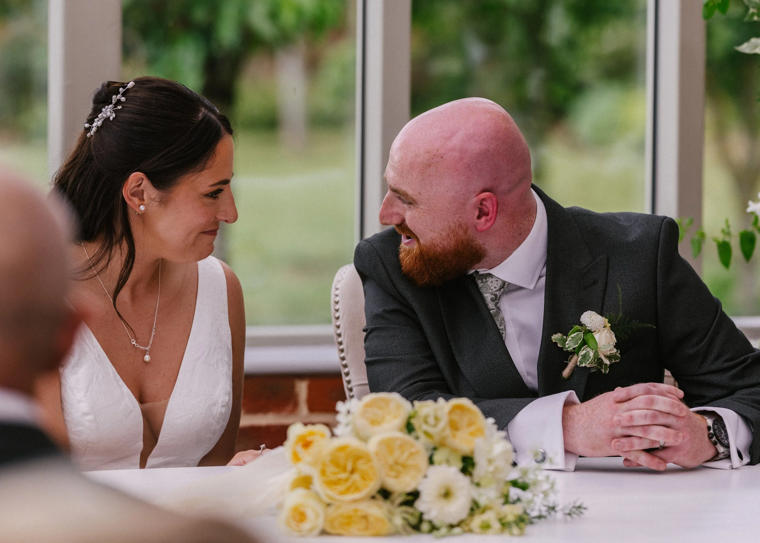 A bride and groom sitting at a table during their wedding. The bride is in a white dress and the groom is in a dark suit with a boutonniere. They are smiling at each other, with a bouquet of yellow roses in front of them and windows with a view of gr