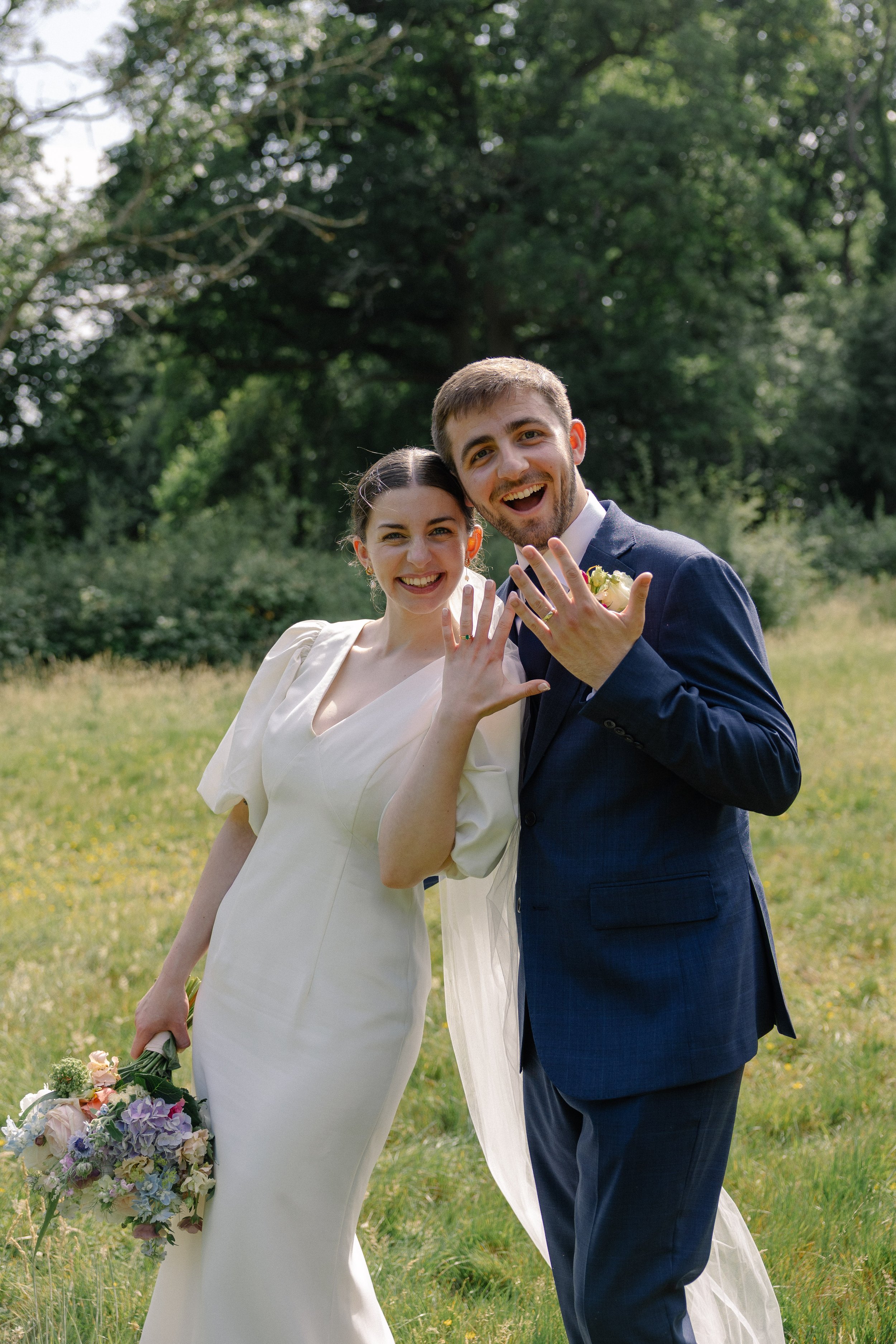 A happy couple, dressed in wedding attire, posing outdoors in a lush green park. The bride is wearing a white dress holding a bouquet of pastel flowers, and the groom is in a dark blue suit showing their wedding rings.
