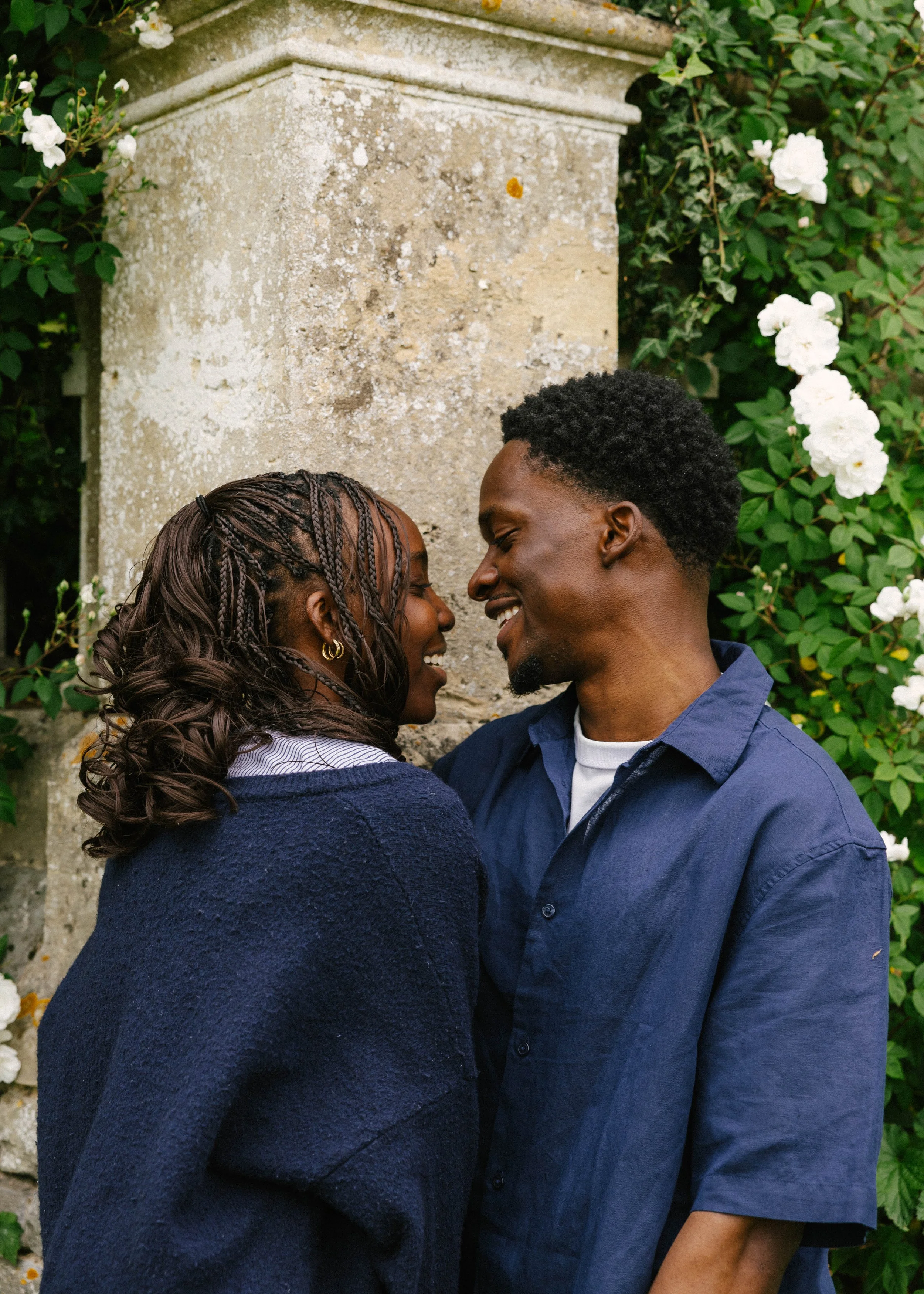 A man and woman standing close together outdoors, smiling and touching foreheads, with green ivy and white flowers in the background.