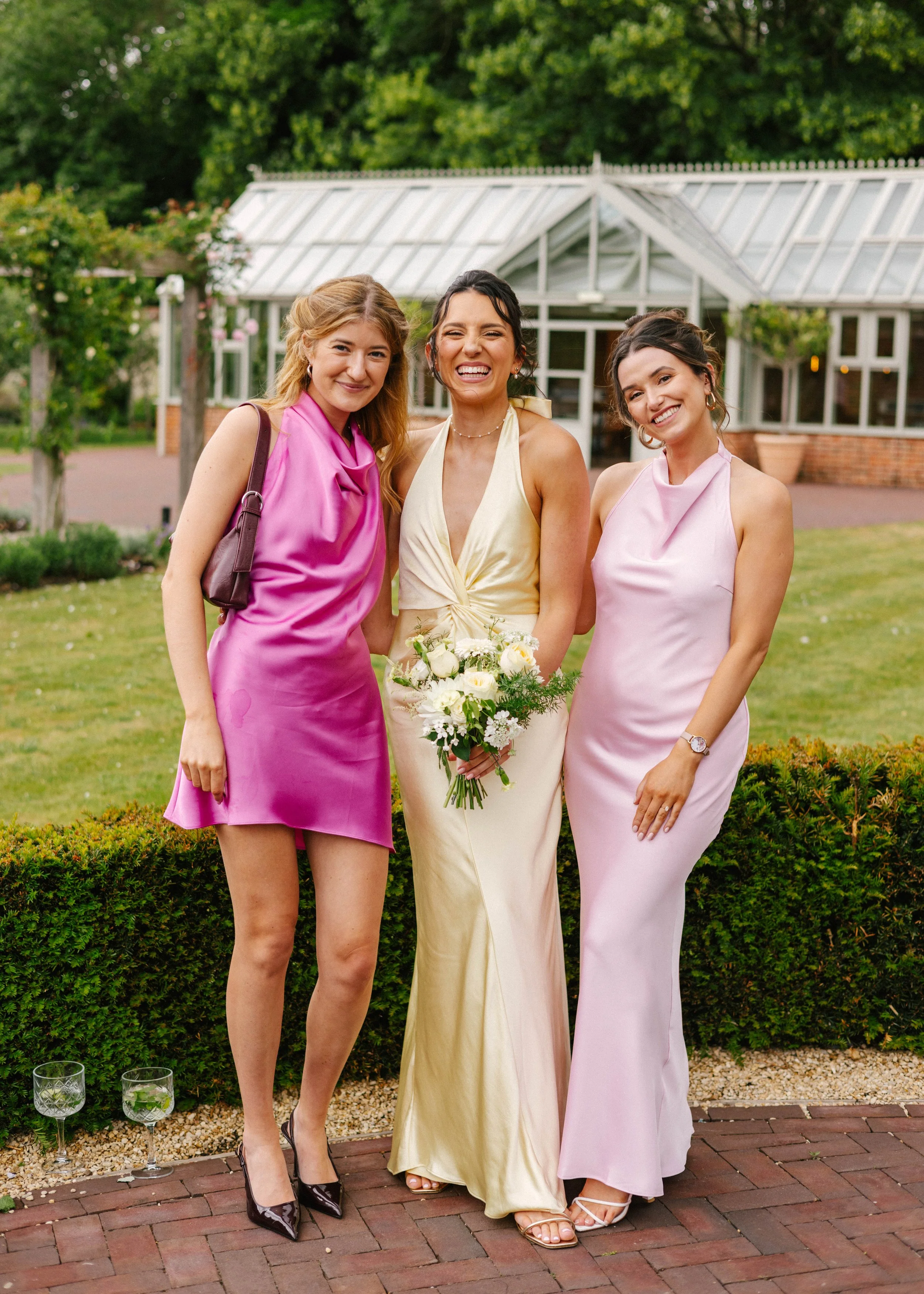 Three women dressed in colorful formal dresses standing outdoors, smiling, with a greenhouse and greenery in the background.