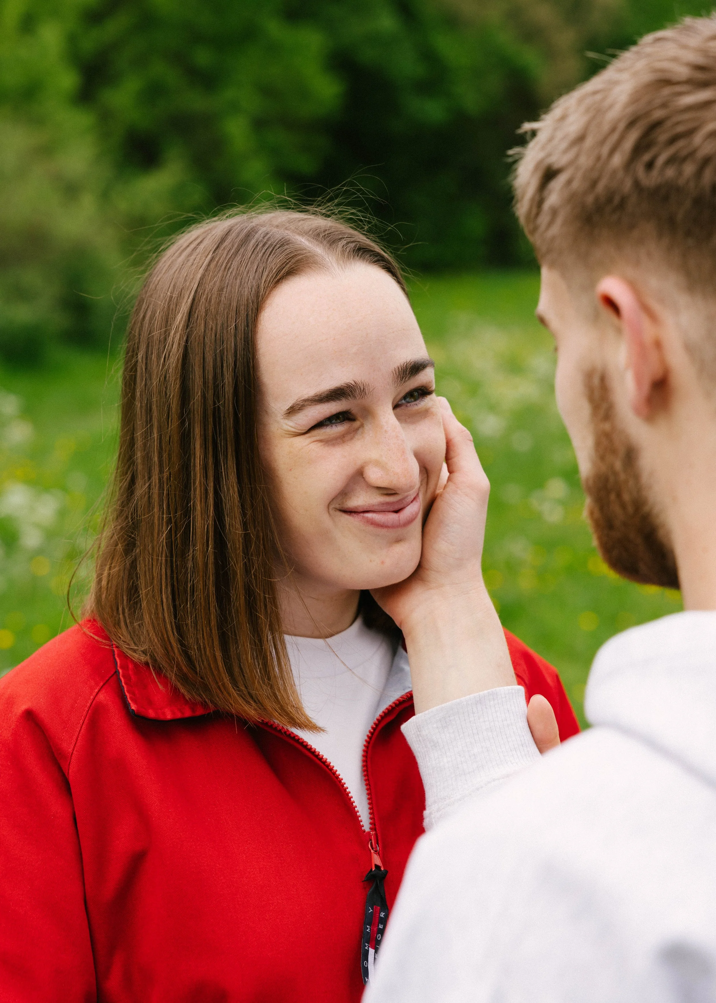 A young woman with long brown hair and light skin smiling at a person in a white hoodie, outdoors with green trees in the background.