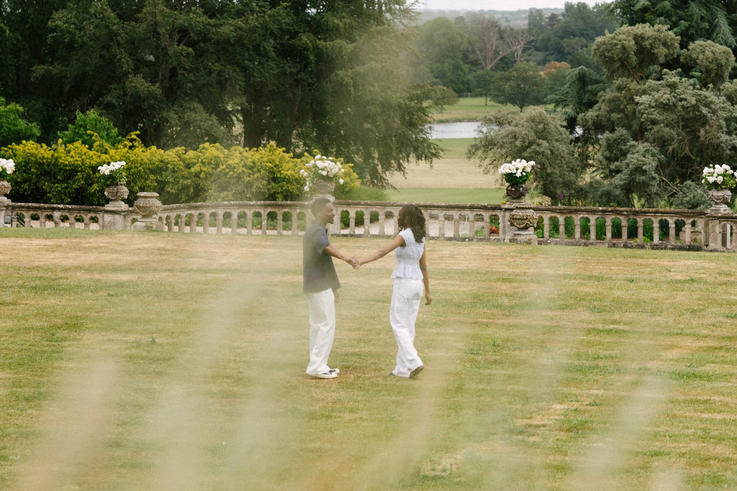 A man and woman holding hands and dancing outdoors on a grassy field with trees and shrubs in the background.