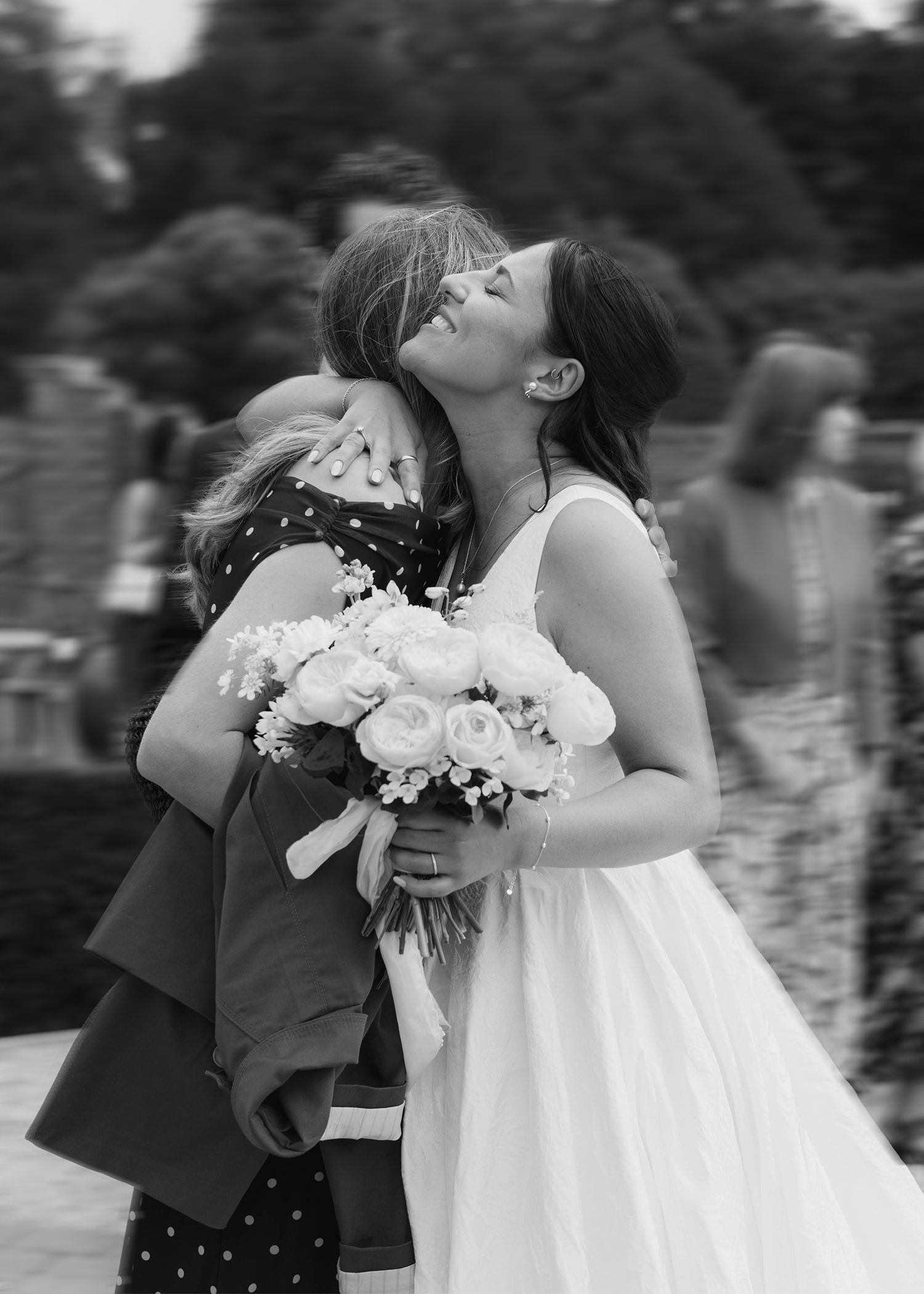 A woman in a wedding dress hugging a child, holding a bouquet of flowers, in an outdoor setting with other people in the background.