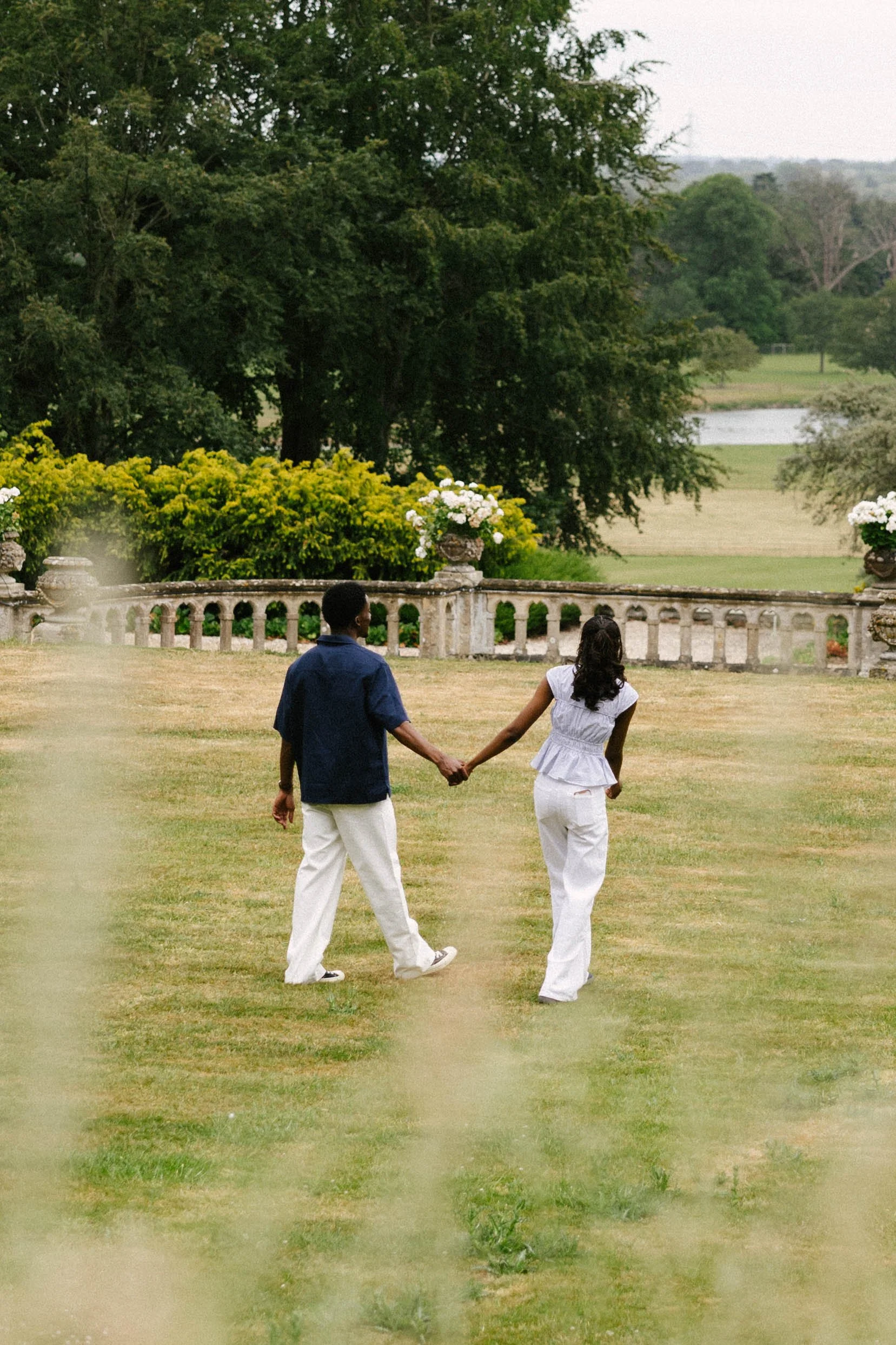 A couple holding hands while walking in a grassy park with trees and a stone railing.