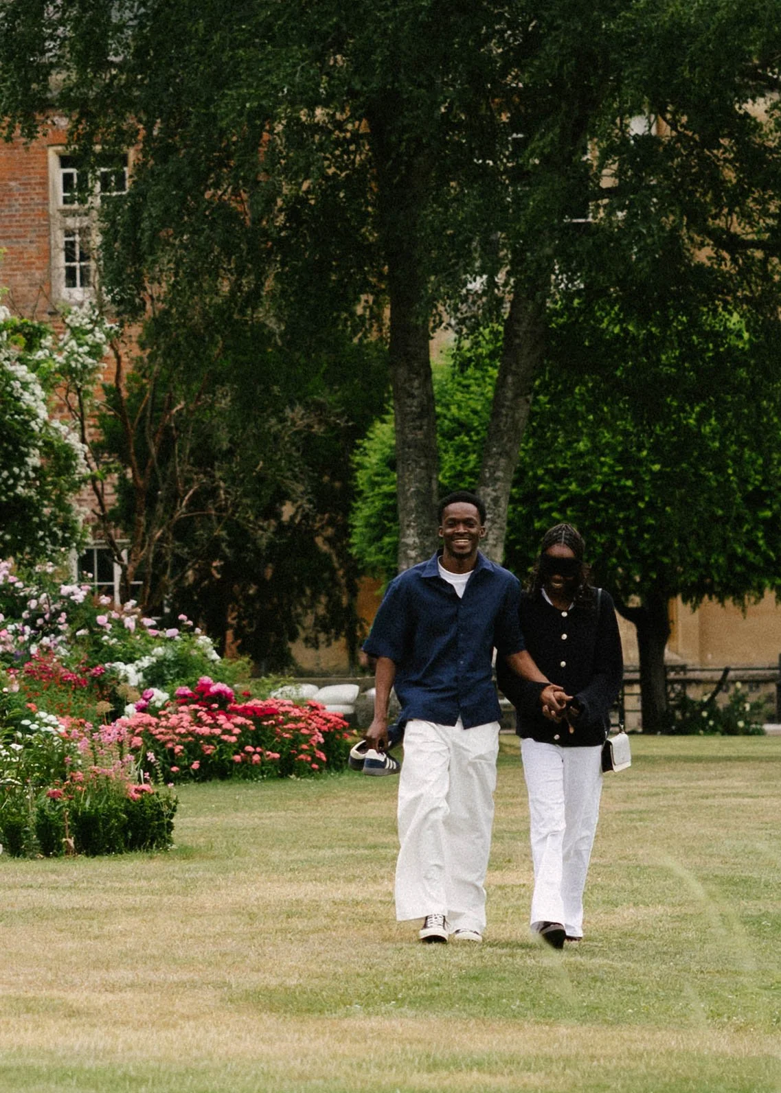 A young man and woman walking and smiling outdoors on a grassy area with flowers and trees, with buildings in the background.