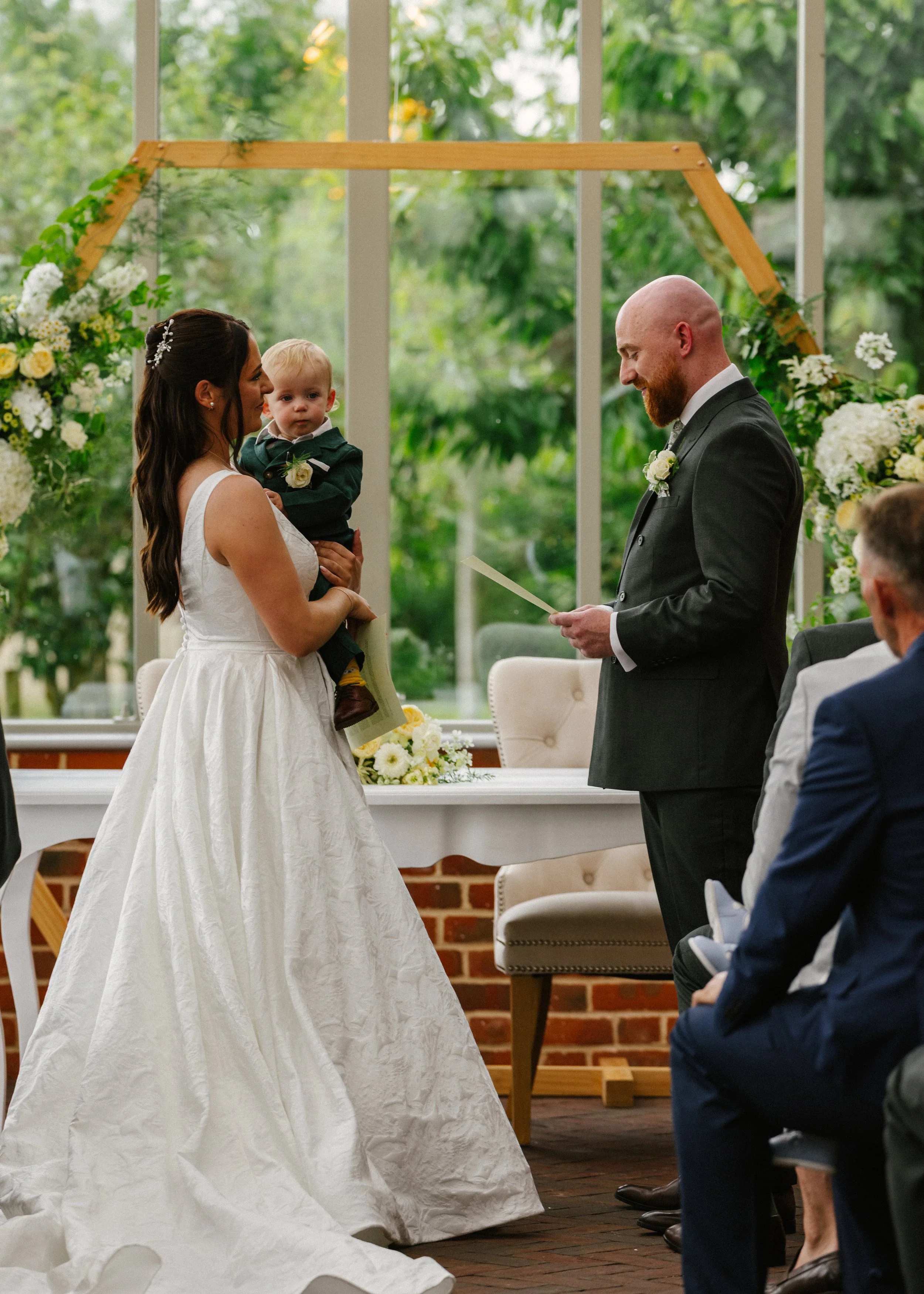Bride and groom exchanging vows at a wedding ceremony while holding a young child, with guests seated nearby and floral decorations in the background.