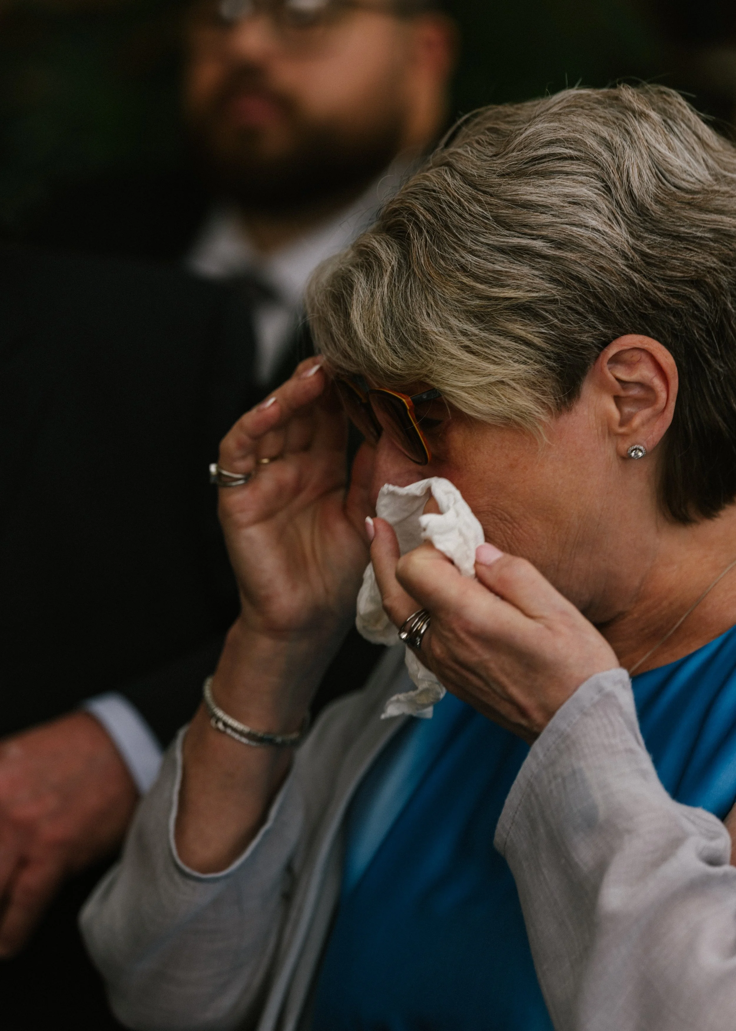 A woman with gray hair wearing glasses and earrings, wiping her eyes with a tissue, being comforted by a person behind her, while another person in the background watches.