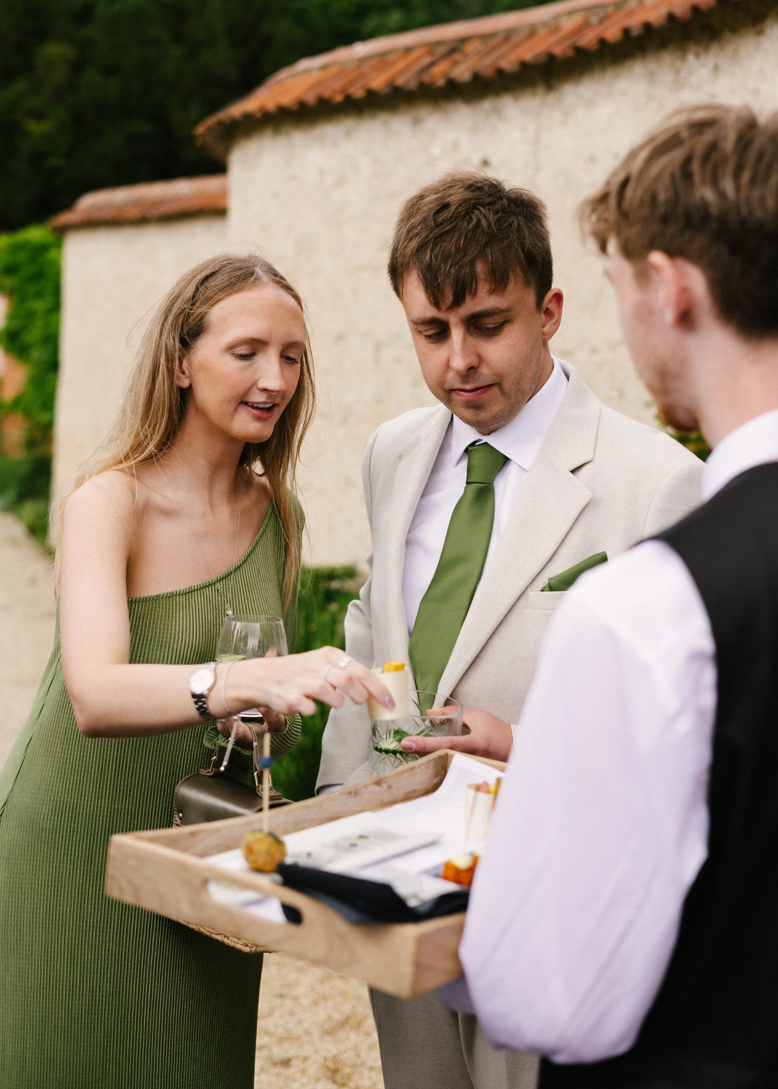 People at an outdoor event, woman in green dress serving food and drinks from a wooden tray to two men, one in a beige suit and the other in black vest, with a building in the background.
