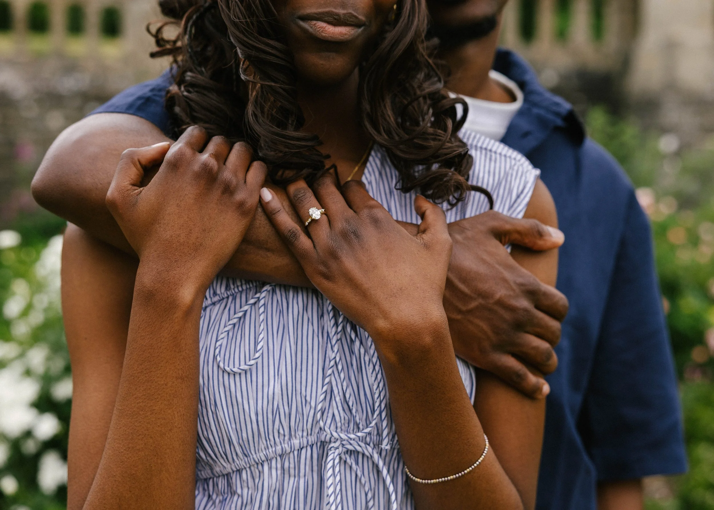 Close-up of two women embracing outdoors. One woman has dark skin and brown curly hair, wearing a striped shirt and an engagement ring. The other woman, partially visible, also has dark skin and is clothed in a navy jacket. The background is blurred 