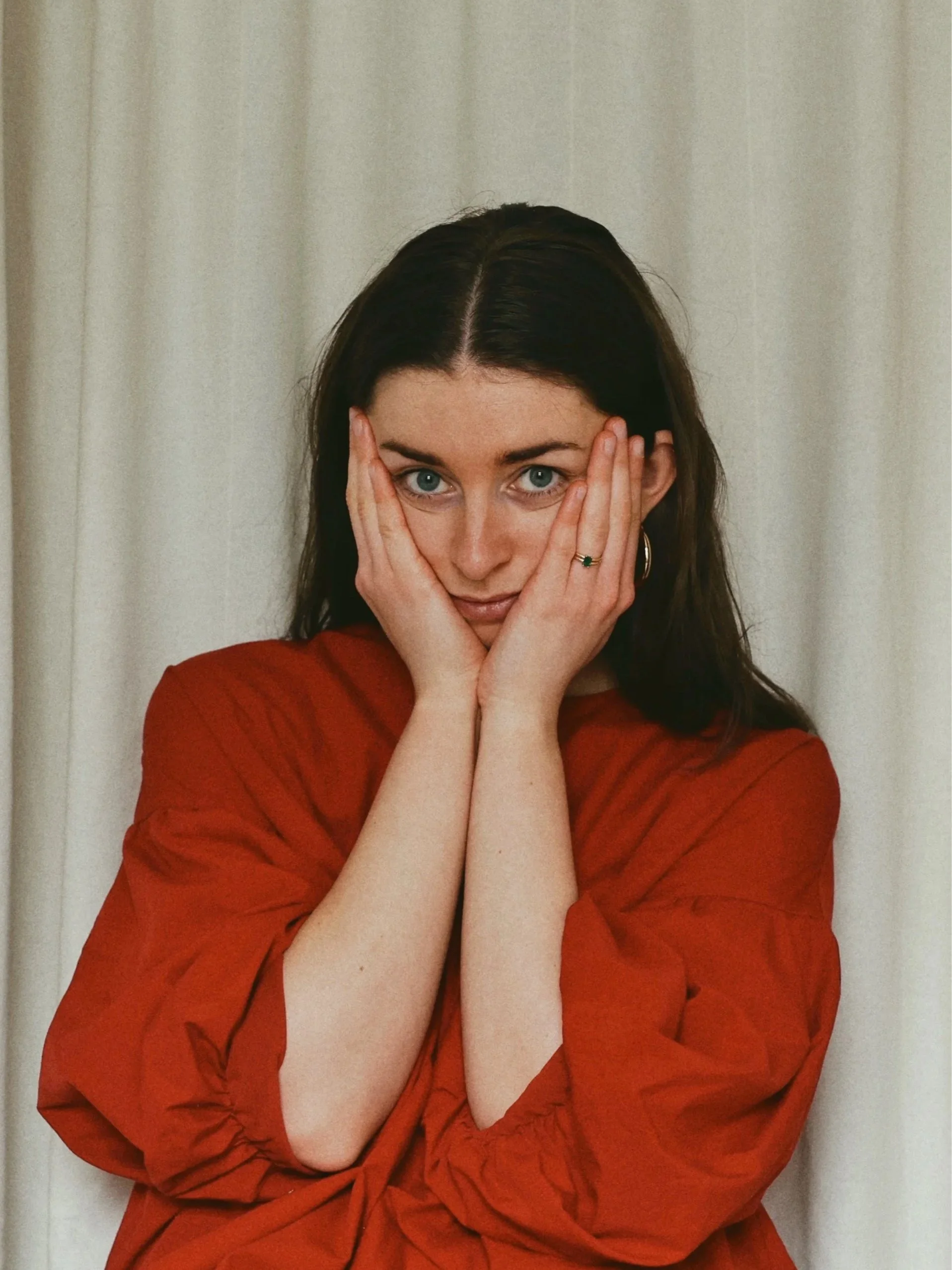 A woman with dark hair and piercing blue eyes, holding her face with both hands, looking directly at the camera, wearing a red top, against a neutral background.
