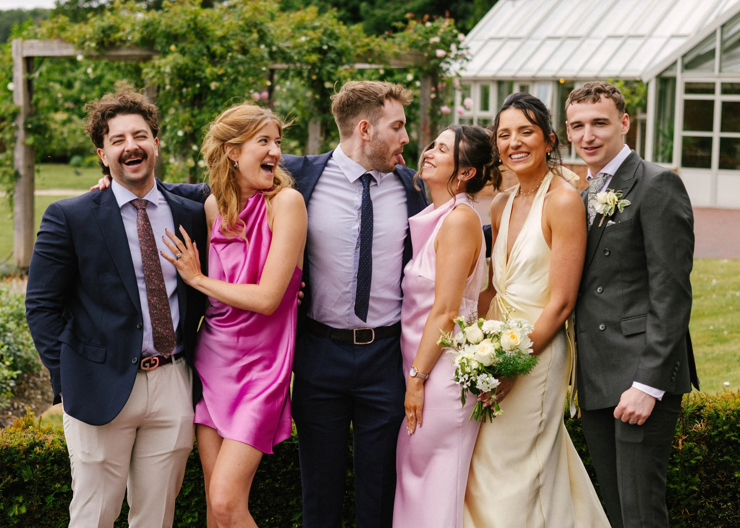 Group of people at a wedding or celebration in a garden, including men in suits and women in colorful dresses, smiling and interacting with each other.
