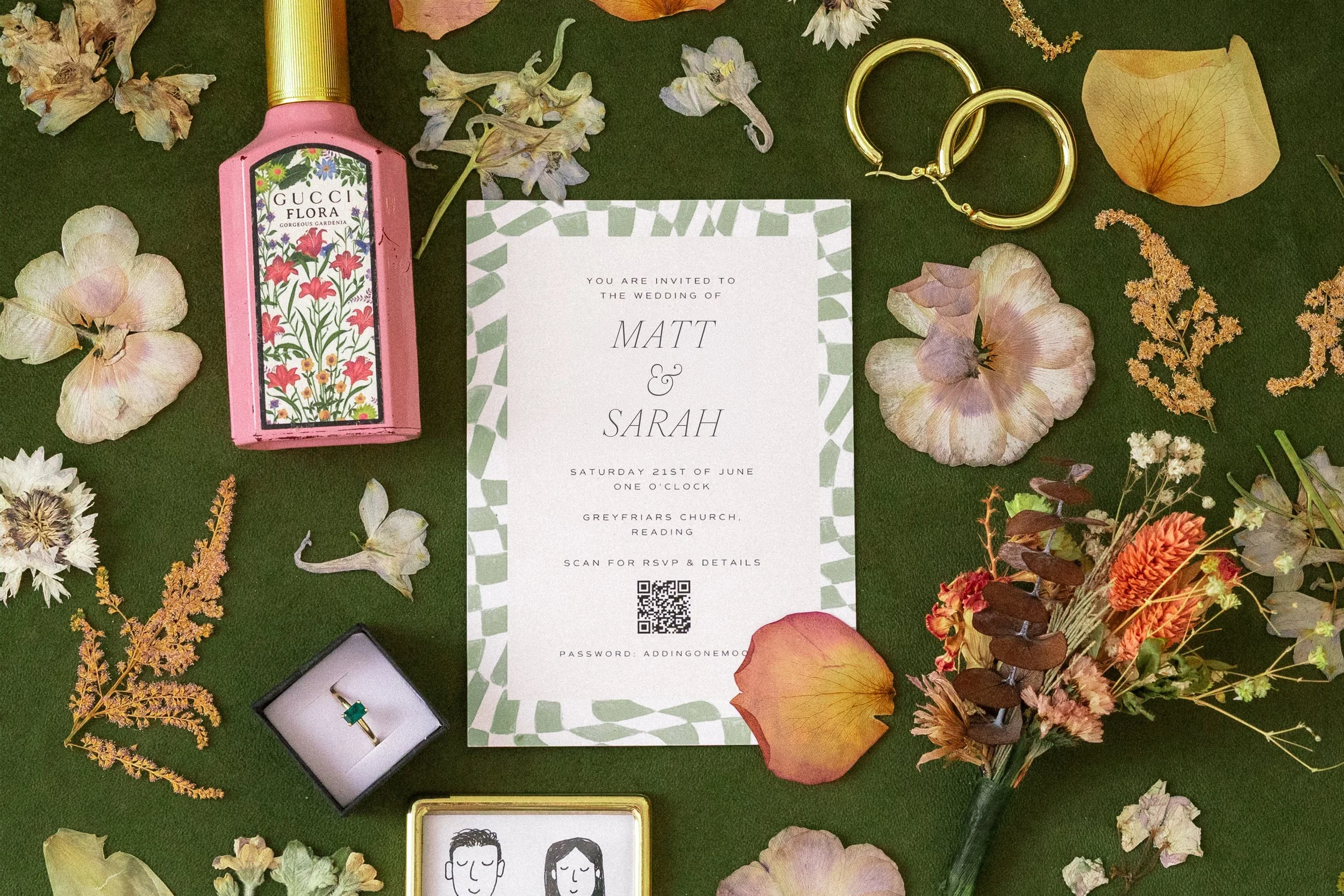 Wedding invitation surrounded by scattered dried flowers, a pink perfume bottle, gold hoop earrings, a ring in a black jewelry box, and a small framed line drawing of a man and woman with closed eyes.
