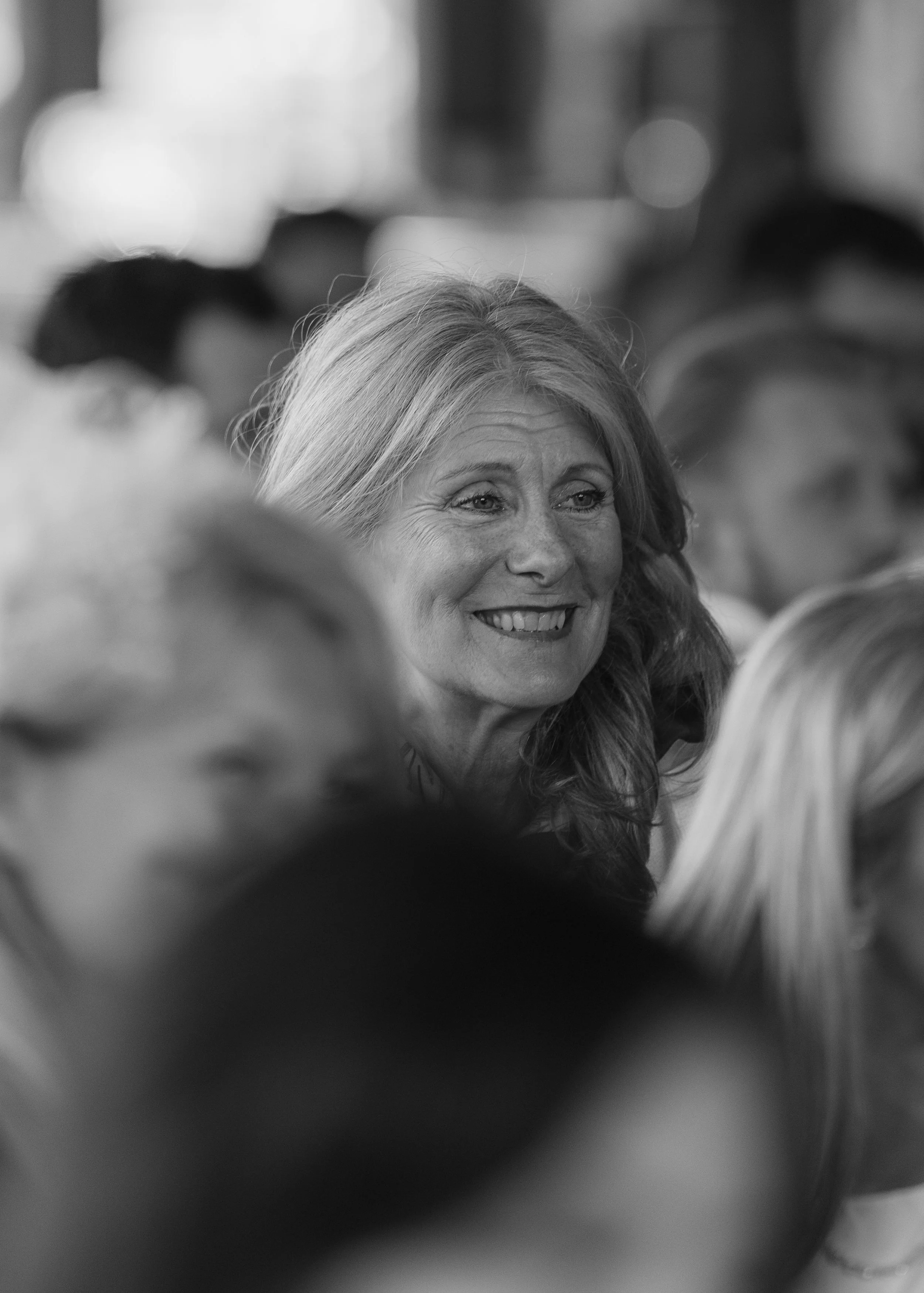 Black and white photo of a smiling woman with long hair, surrounded by other people in an indoor setting.