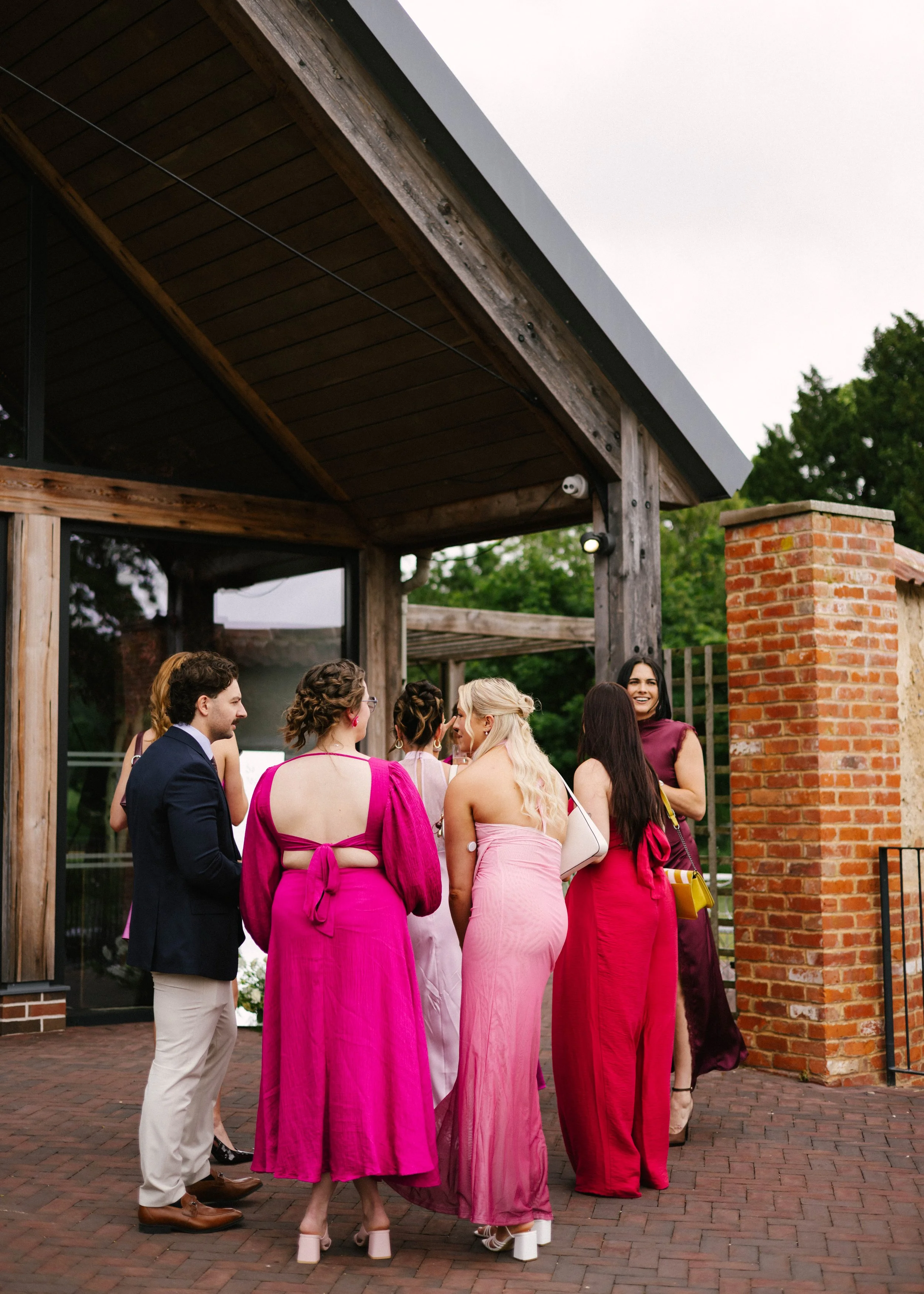 Group of people dressed in colorful formal attire talking outside under a wooden structure with brick pillars.