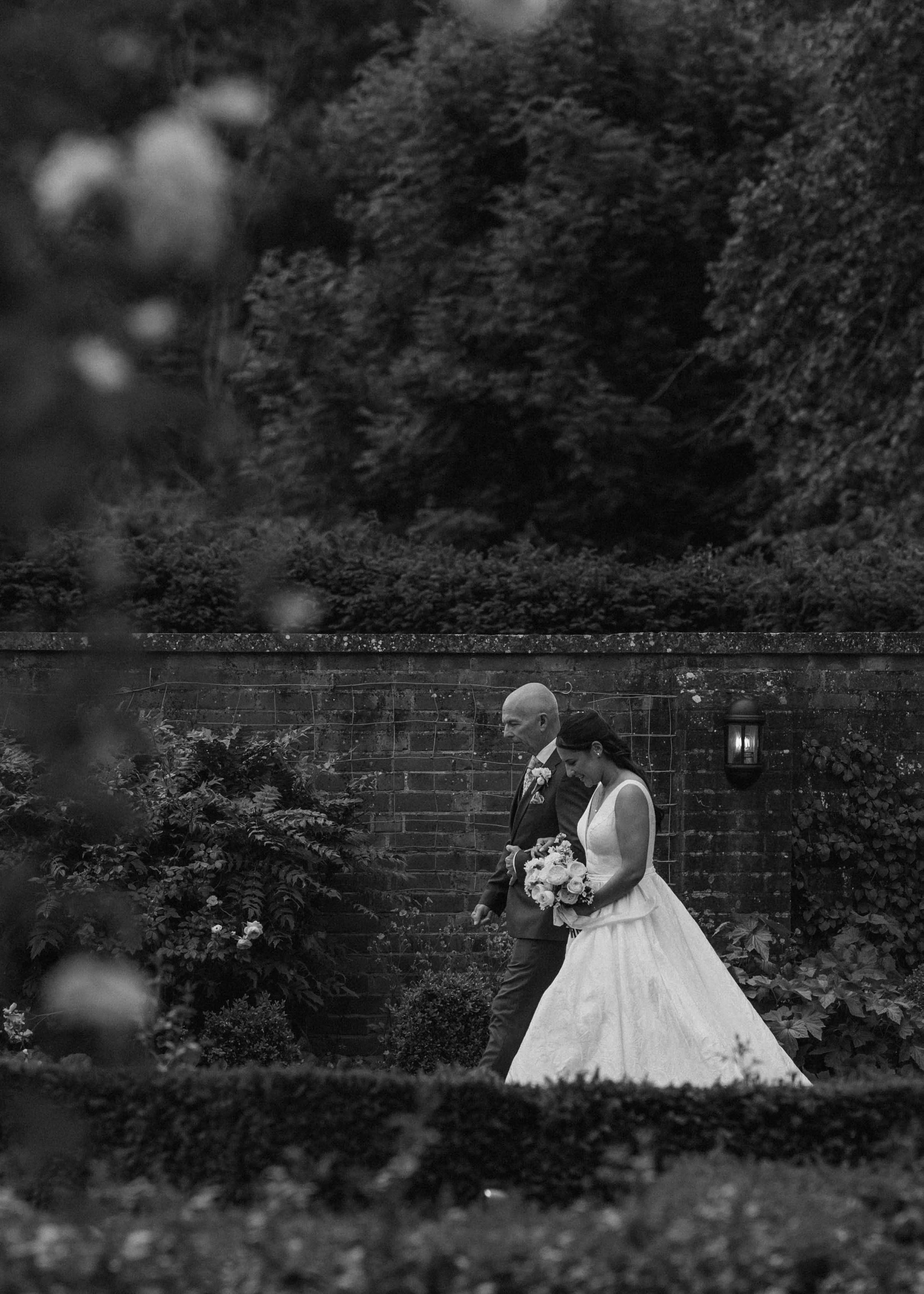 A bride and groom walking together outdoors in a garden setting, with the bride holding a bouquet of flowers, both dressed in formal wedding attire.