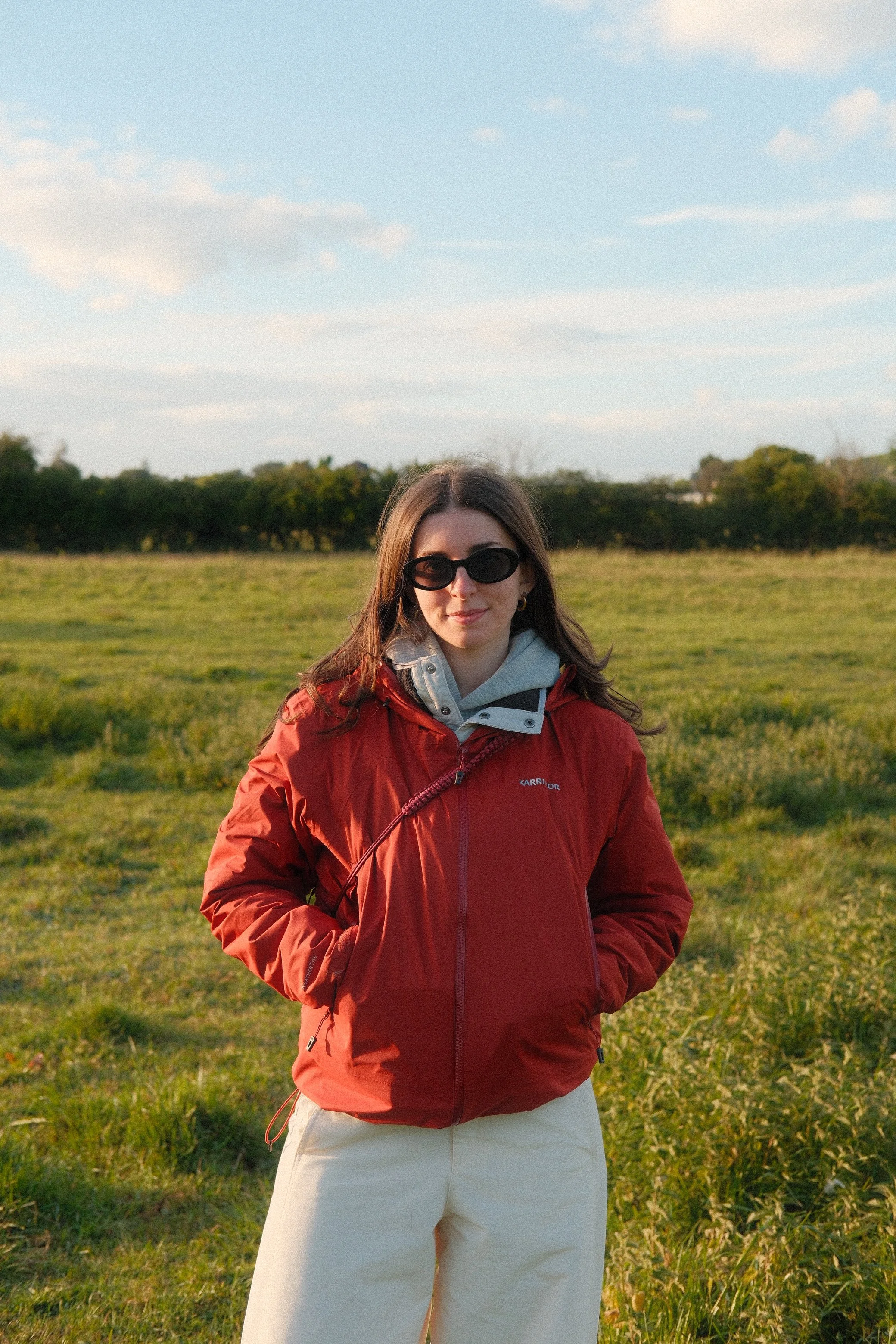 A woman wearing a red jacket, white hoodie, and white pants standing outdoors with a green field and trees in the background, under a partly cloudy sky.
