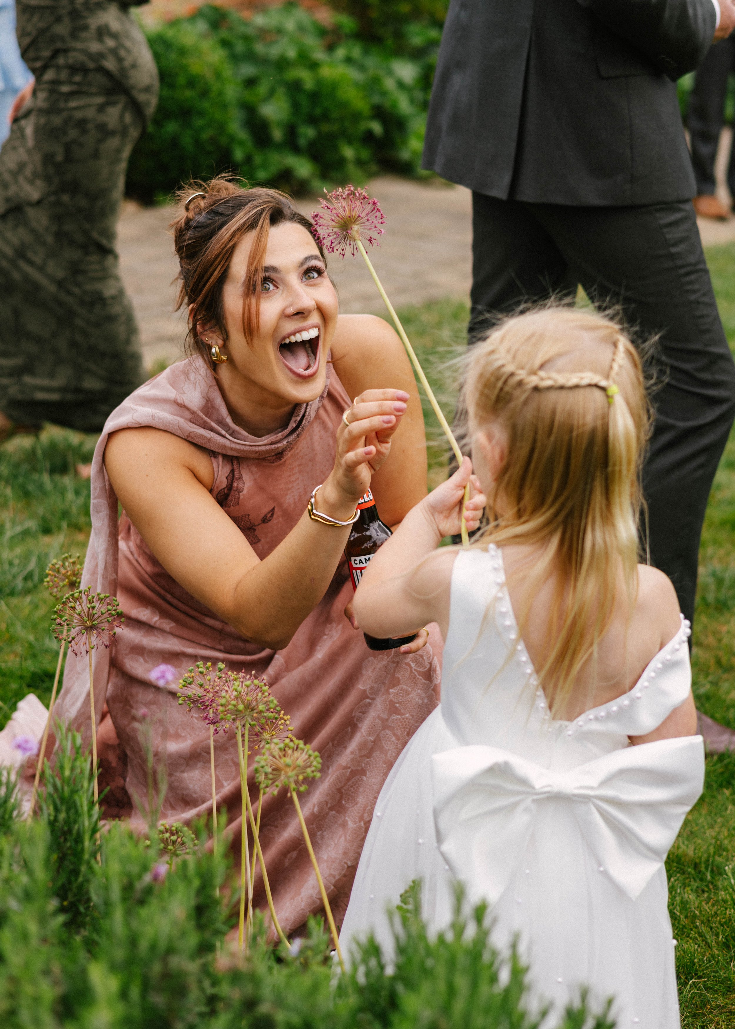A woman with short brown hair, wearing gold earrings, and a pink dress with lace, happily interacts with a young girl with blonde hair, who is in a white dress, in a garden with green grass and flowers. The woman is holding a flower and appears to be