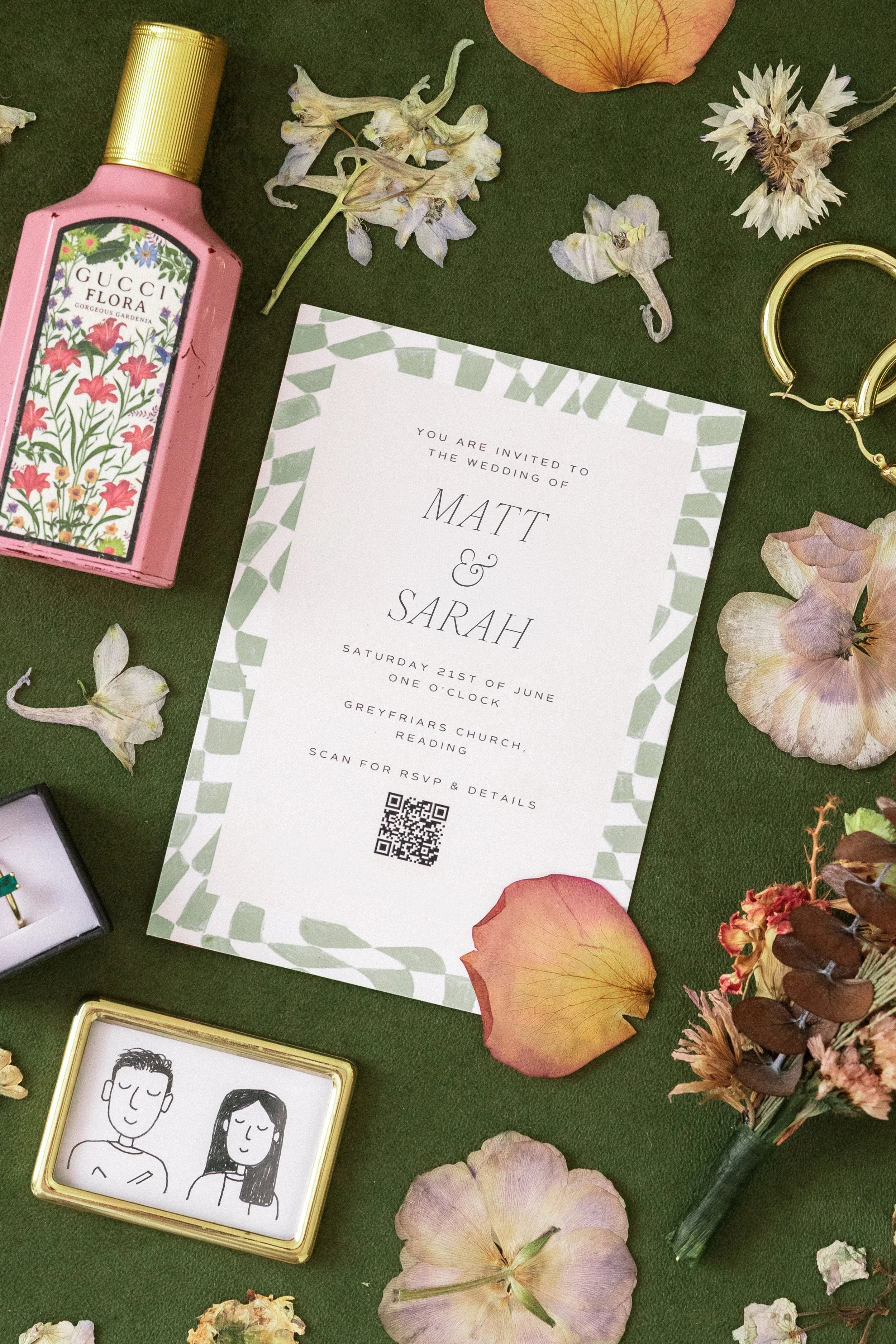 A wedding invitation card for Matt and Sarah, with floral border, surrounded by dried flower petals, a Gucci Flora perfume bottle, a pair of gold earrings, and a small illustration of a man and woman, all arranged on a green surface.