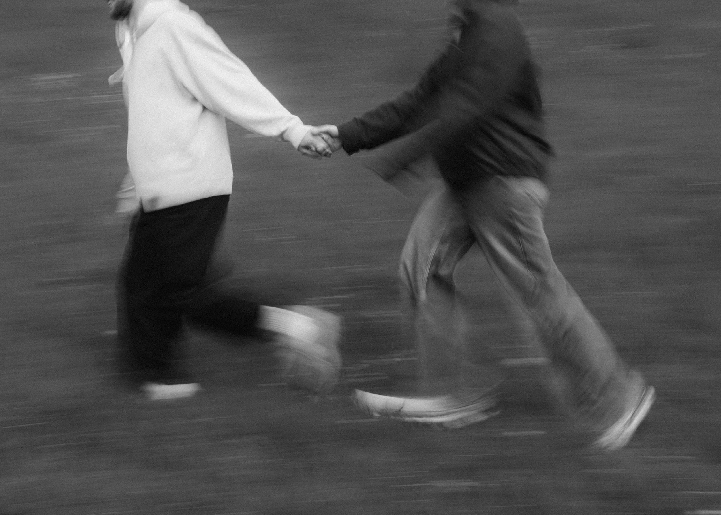 Two people are holding hands while walking or running. The photo is in black and white and has a motion blur effect.