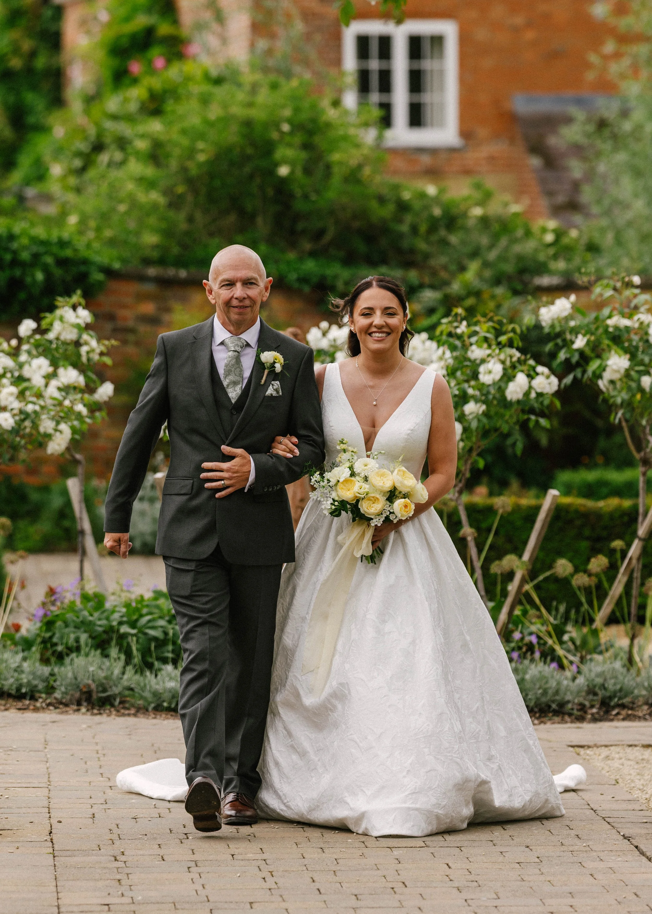 A bride and an older man, possibly her father, walking down an outdoor wedding aisle. The bride is holding a bouquet of white and yellow roses and is wearing a white wedding gown. The man is in a dark gray suit with a boutonniere. They are smiling, s