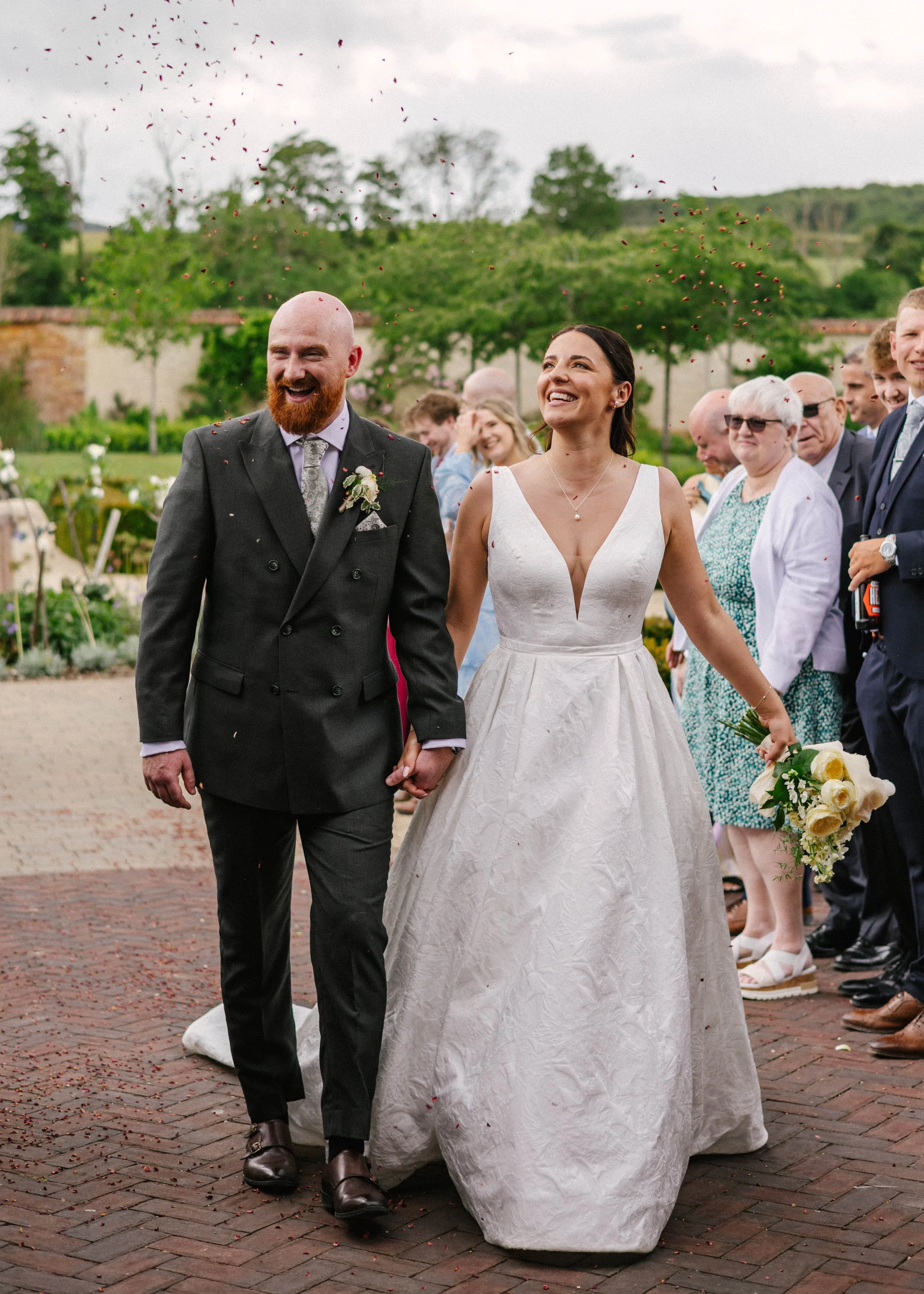 A newlywed couple walking hand in hand outdoors during their wedding, surrounded by friends and family, with flower petals falling around them.