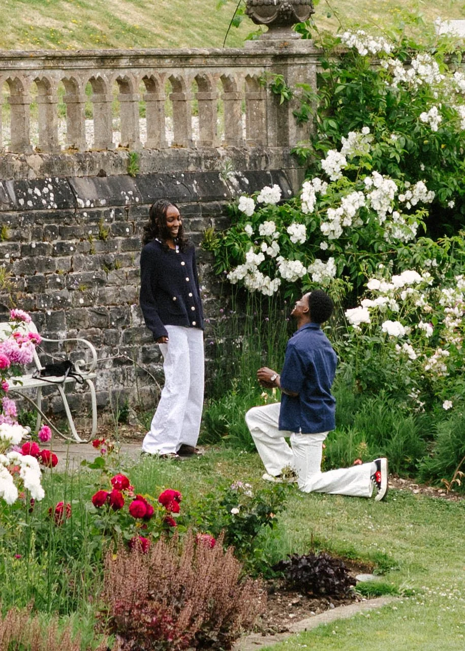 A man proposing marriage to a woman in a garden with colorful flowers and a stone wall.