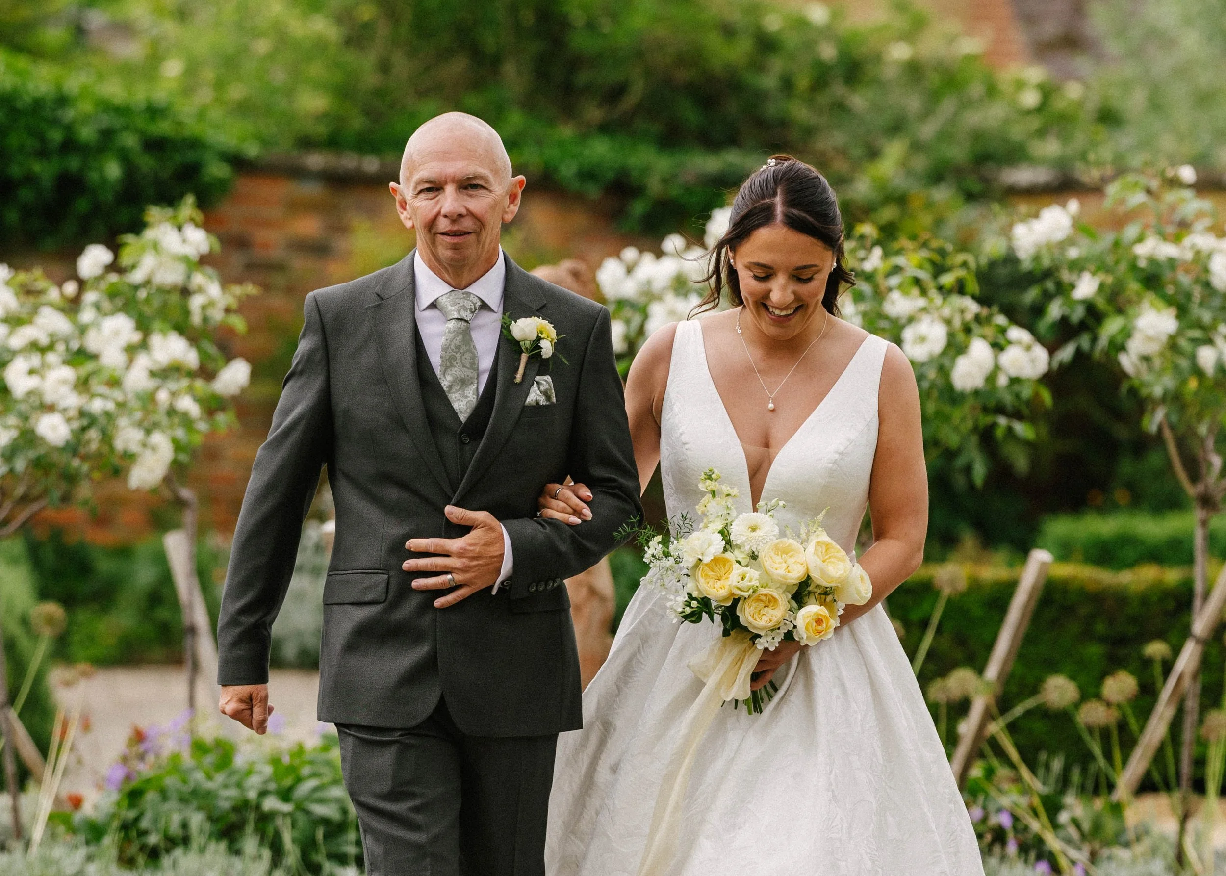 A bride and an older man walking together outdoors at a wedding, with greenery and white flowers in the background. The bride is holding a bouquet of white and yellow flowers and smiling, while the man, dressed in a suit with a boutonniere, is walkin