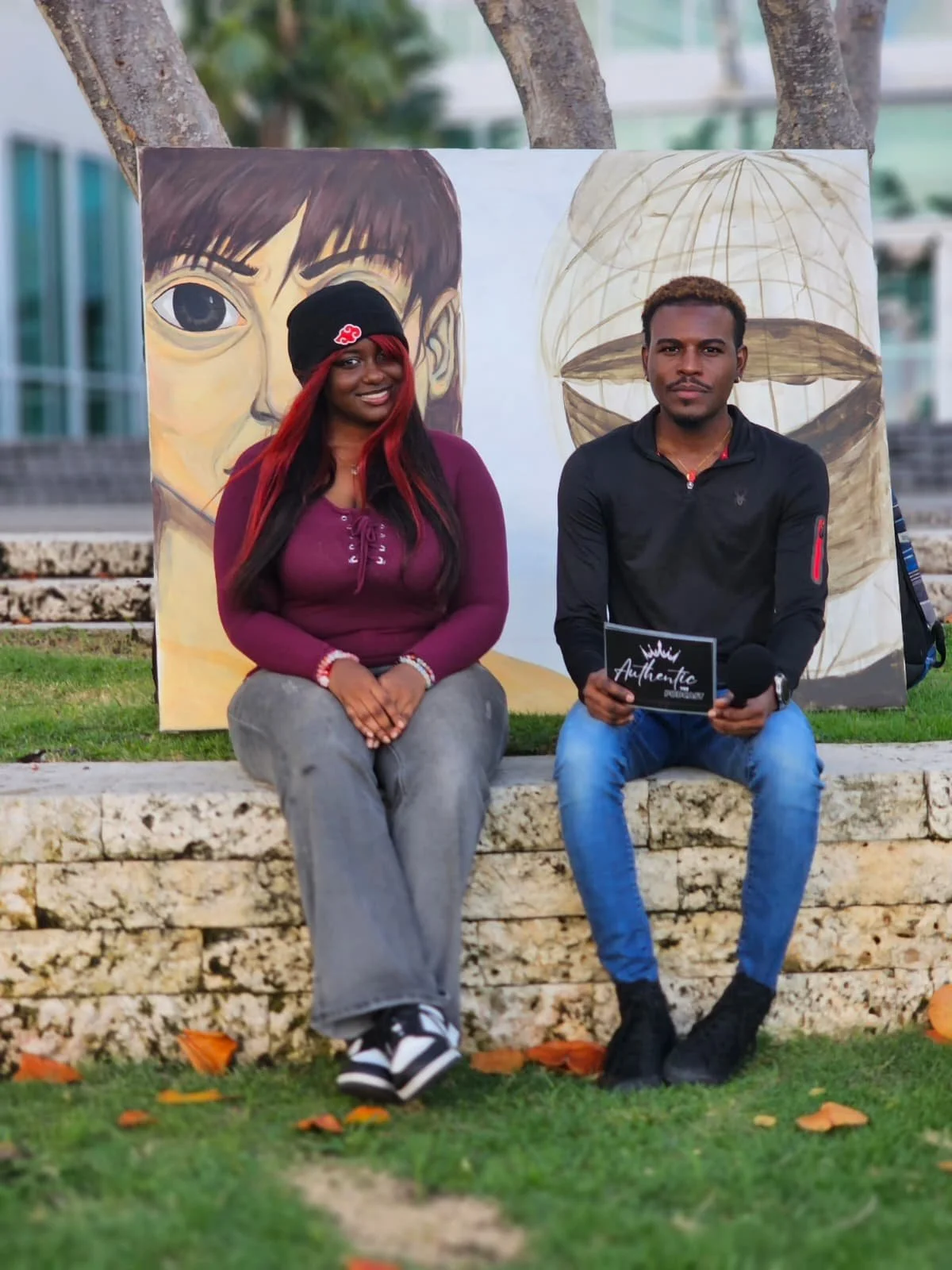 A man and woman sitting on a stone bench outdoors, with colorful paintings behind them. The woman has long red and black hair, wears a black beanie with a red logo, a purple hoodie, gray pants, and sneakers. The man has short dark hair, wears a black