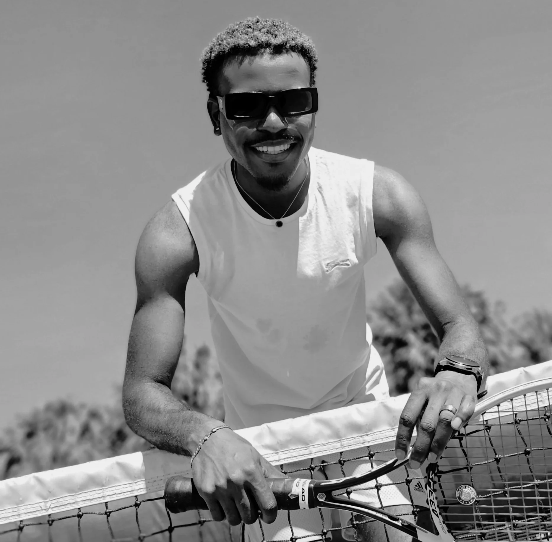 A person with short, curly hair wearing sunglasses and a white sleeveless shirt, smiling while riding a bicycle in an outdoor setting, with a fence in the foreground.