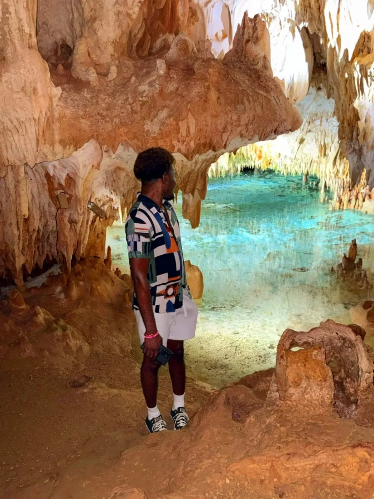 A man standing inside a cave with colorful rock formations and a body of turquoise water, looking towards the water.